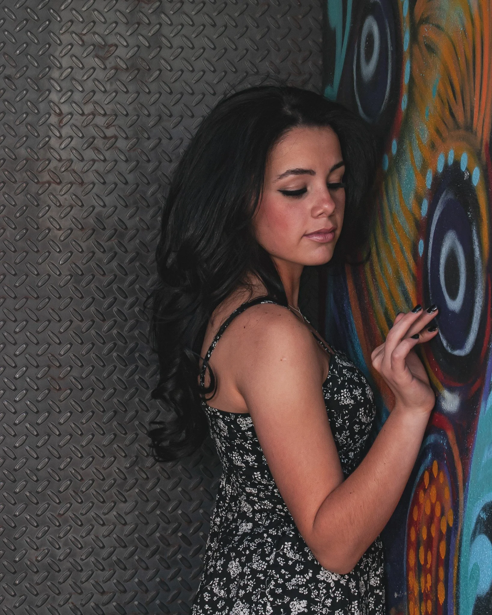 A woman with long black hair and a floral dress standing beside a colorful painted wall. The wall features abstract designs with circles and vibrant colors.