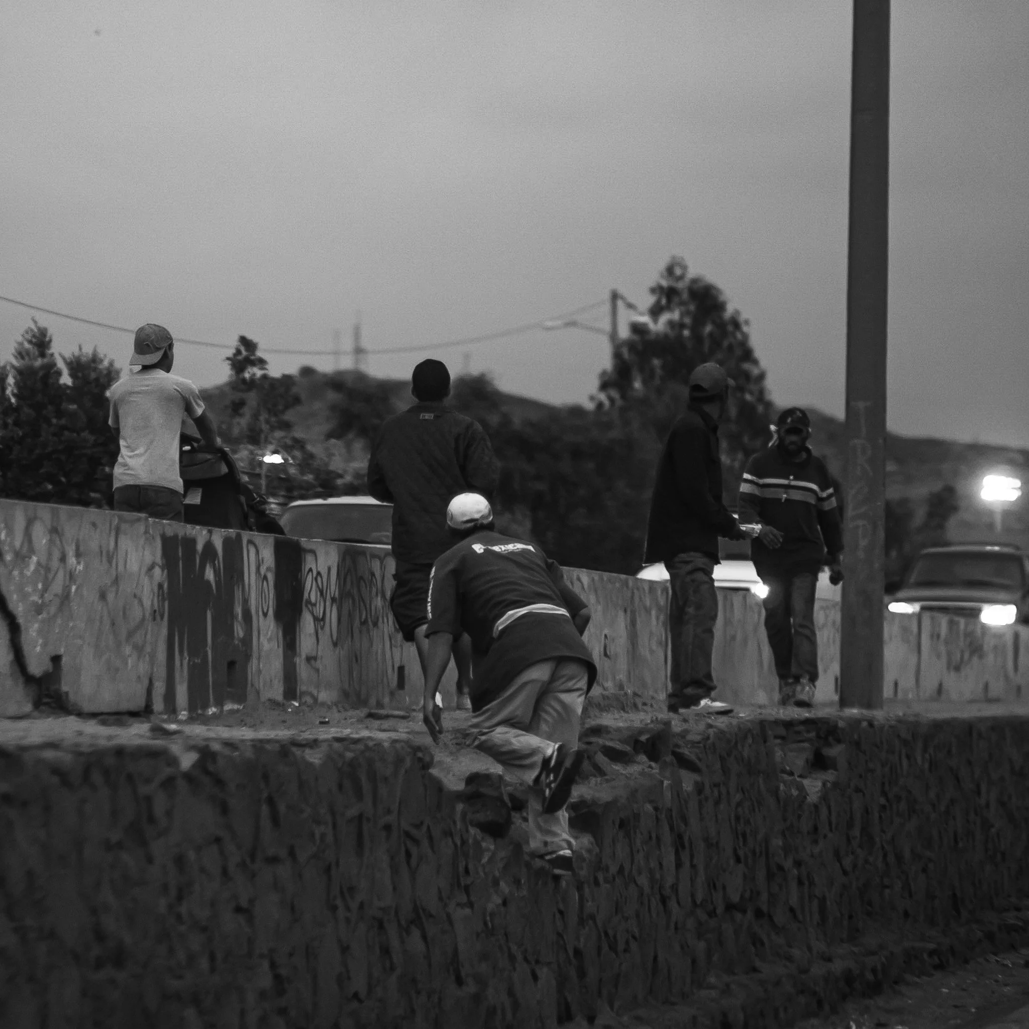 Group of five young men standing and one climbing a stone ledge alongside a graffiti-covered wall, with cars visible in the background.