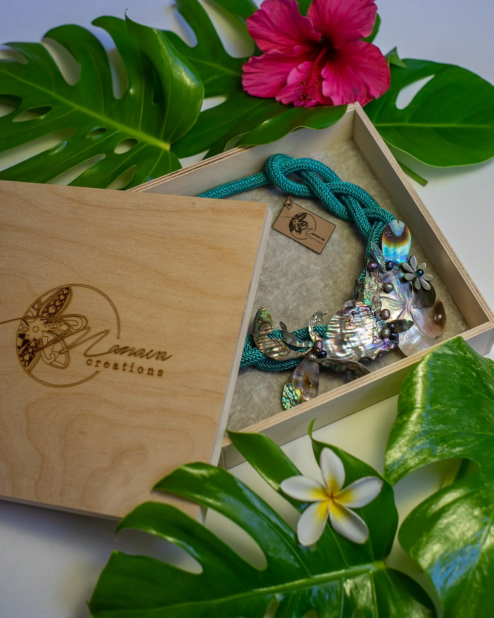 A wooden box with a necklace inside, surrounded by green tropical leaves, pink hibiscus flower, and white plumeria flower.