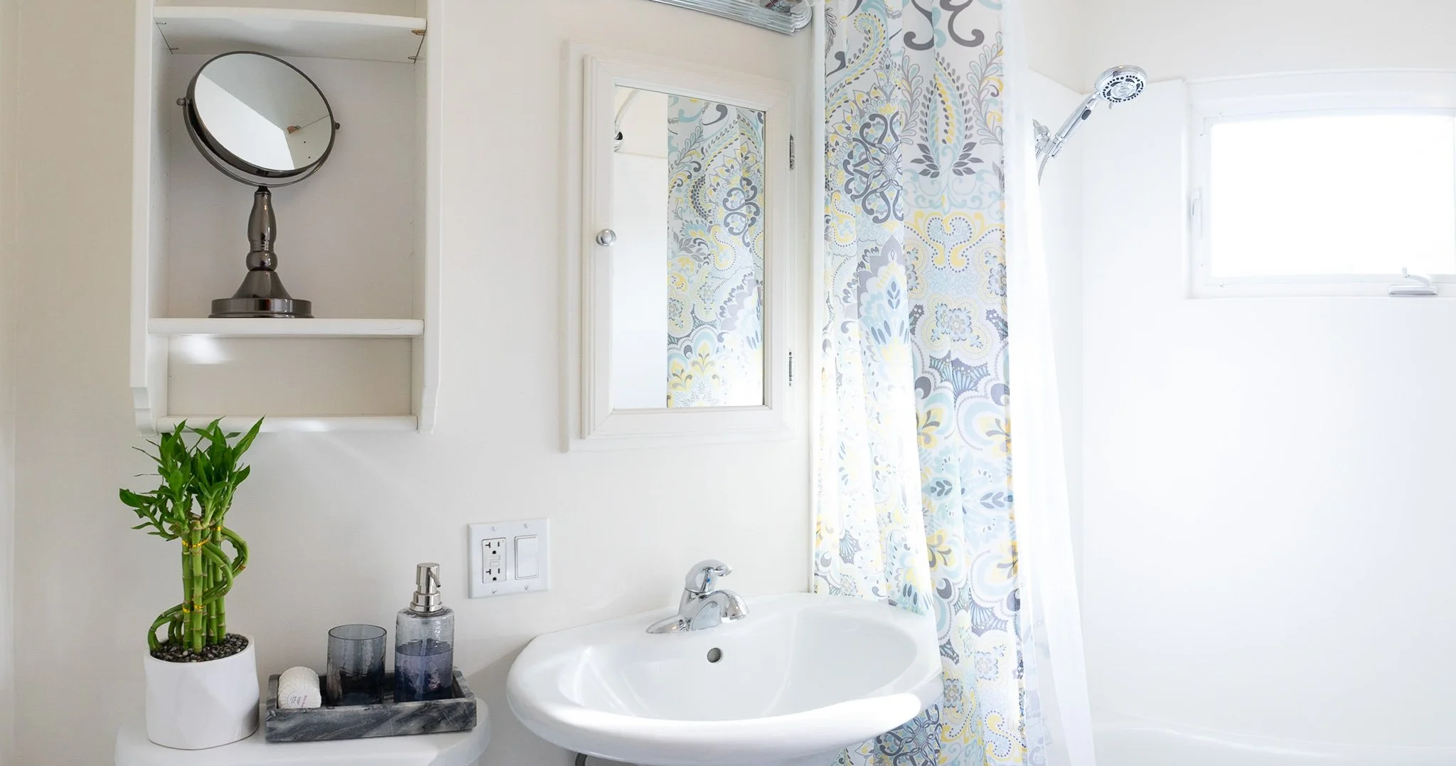 A small bathroom with a white sink, a mirror, a shower curtain with a floral pattern, a small potted bamboo plant, and a soap dispenser on a marble tray.