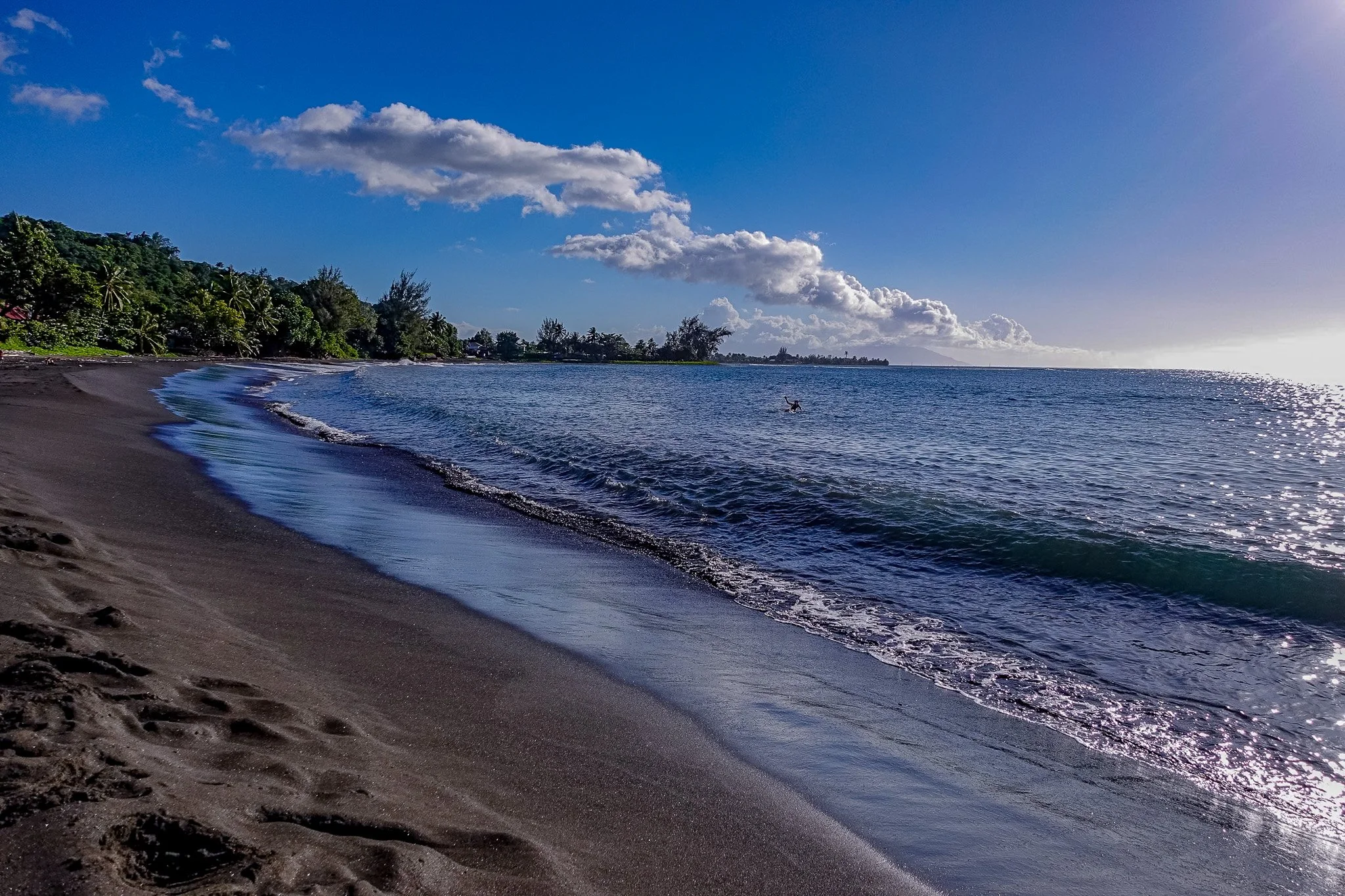 Sunset view of a beach, with calm ocean waves, dark sand, and green trees in the background under a partly cloudy sky.