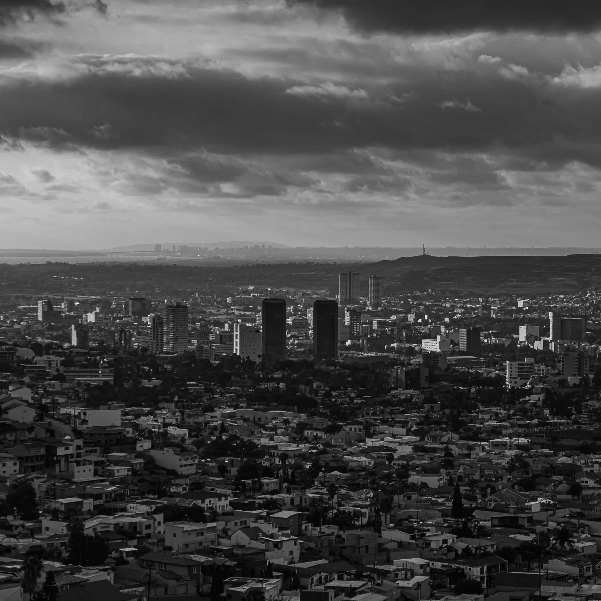 Black and white aerial view of a city skyline with tall buildings, residential houses, and a cloudy sky.