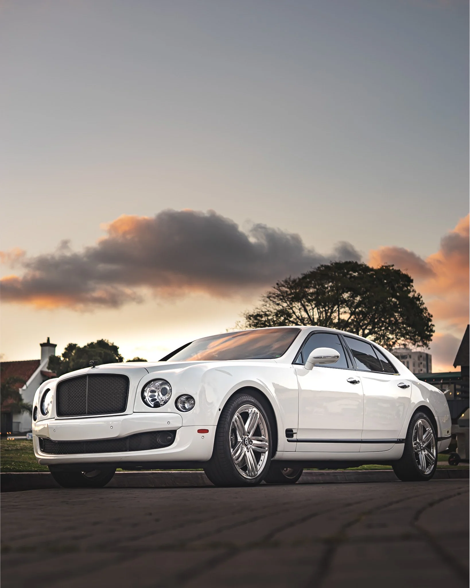 A white luxury sedan parked outdoors during sunset with trees and suburban houses in the background.