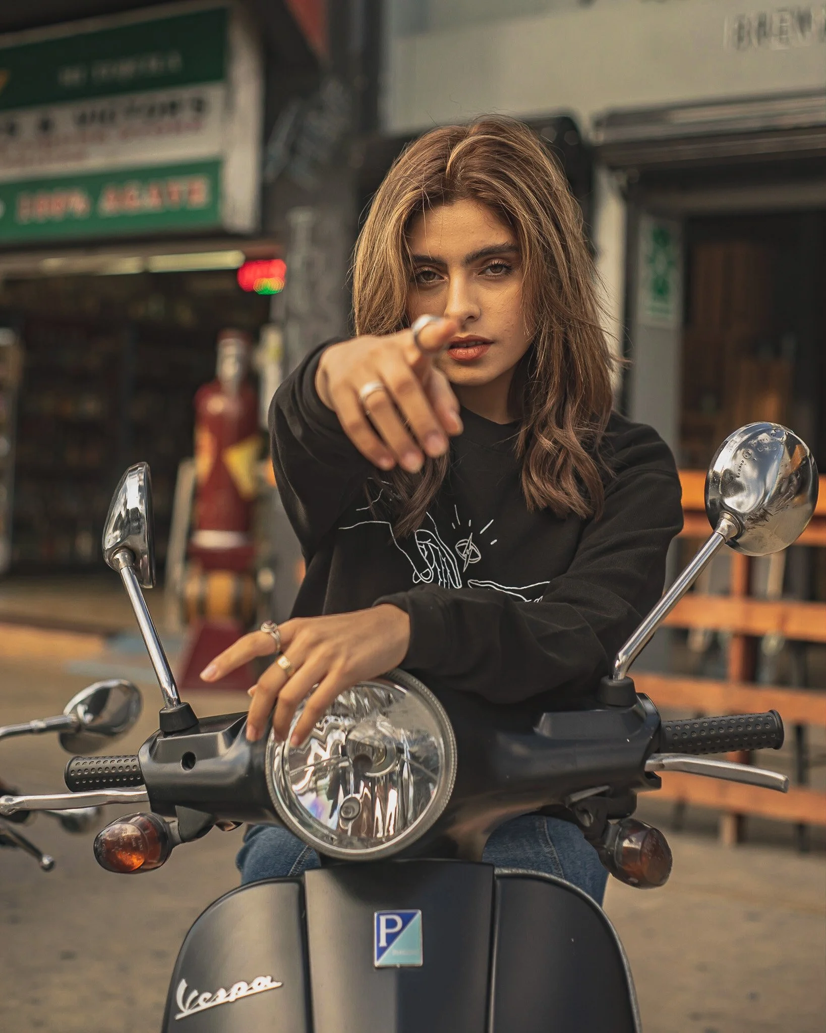 A young woman with shoulder-length brown hair and a black shirt sitting on a Vespa scooter, pointing directly at the camera.