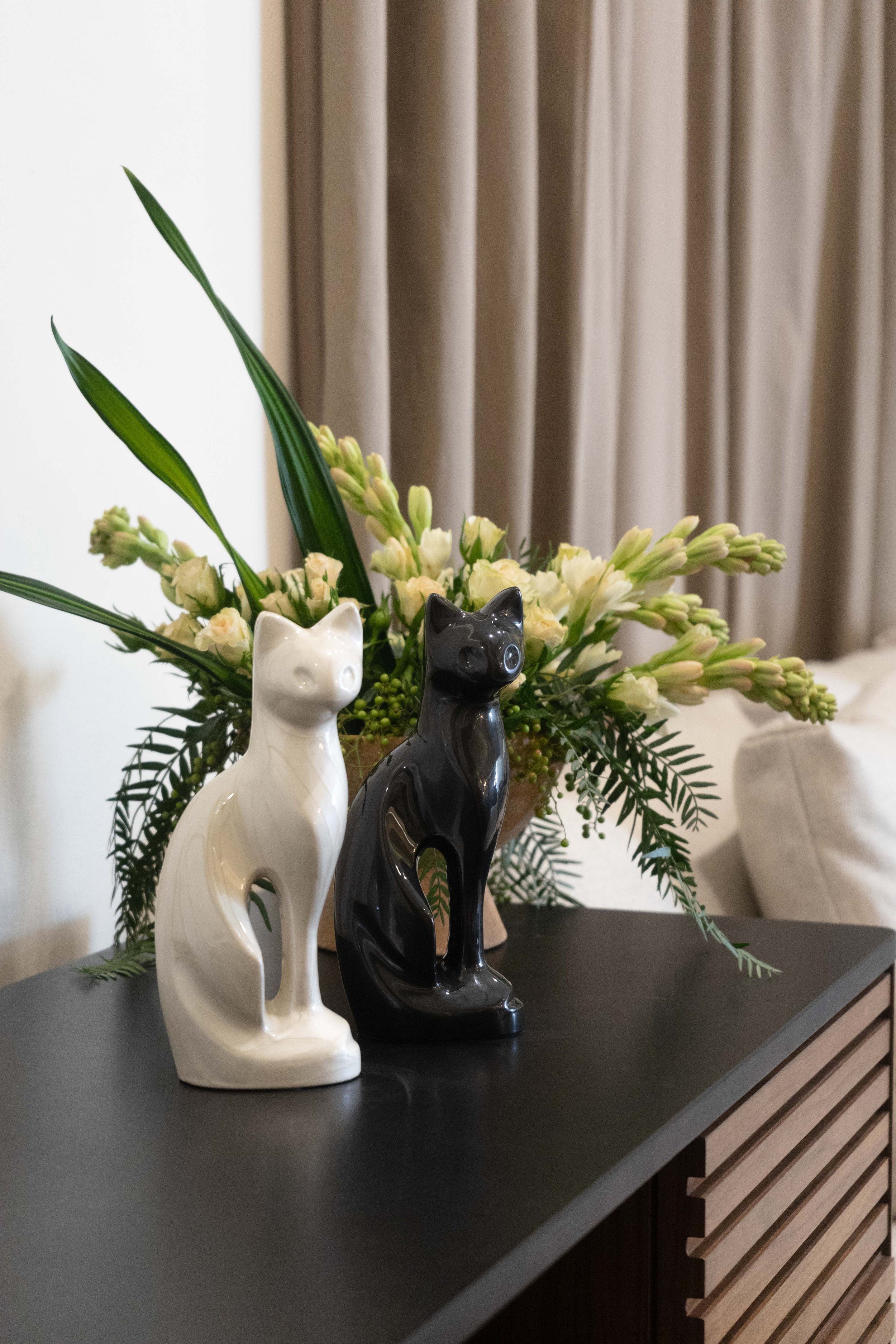 Ceramic cat figurines, one white and one black, placed on a black surface with a floral arrangement in the background.