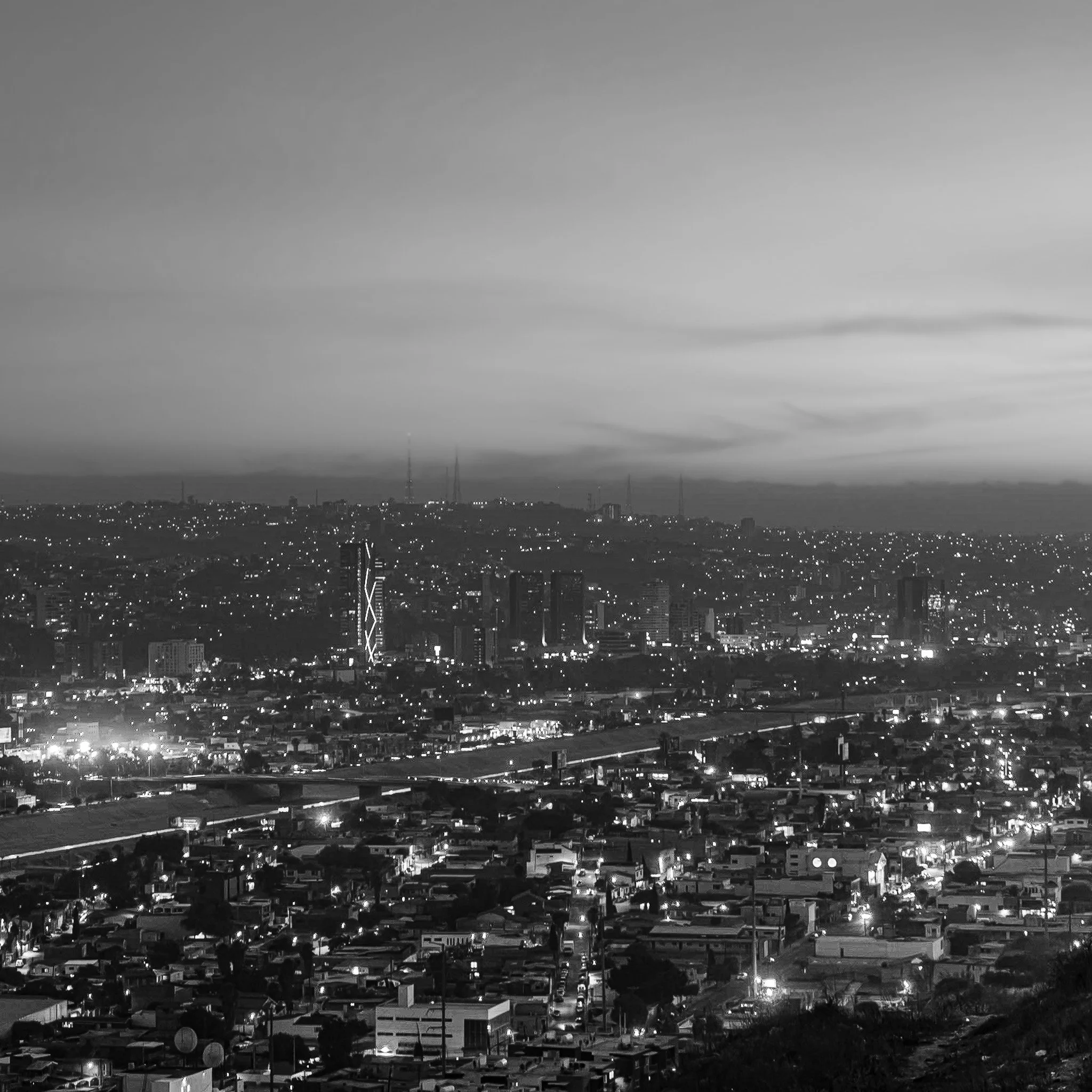 Night view of a city skyline with high-rise buildings, illuminated streets, and a vast urban landscape in black and white.