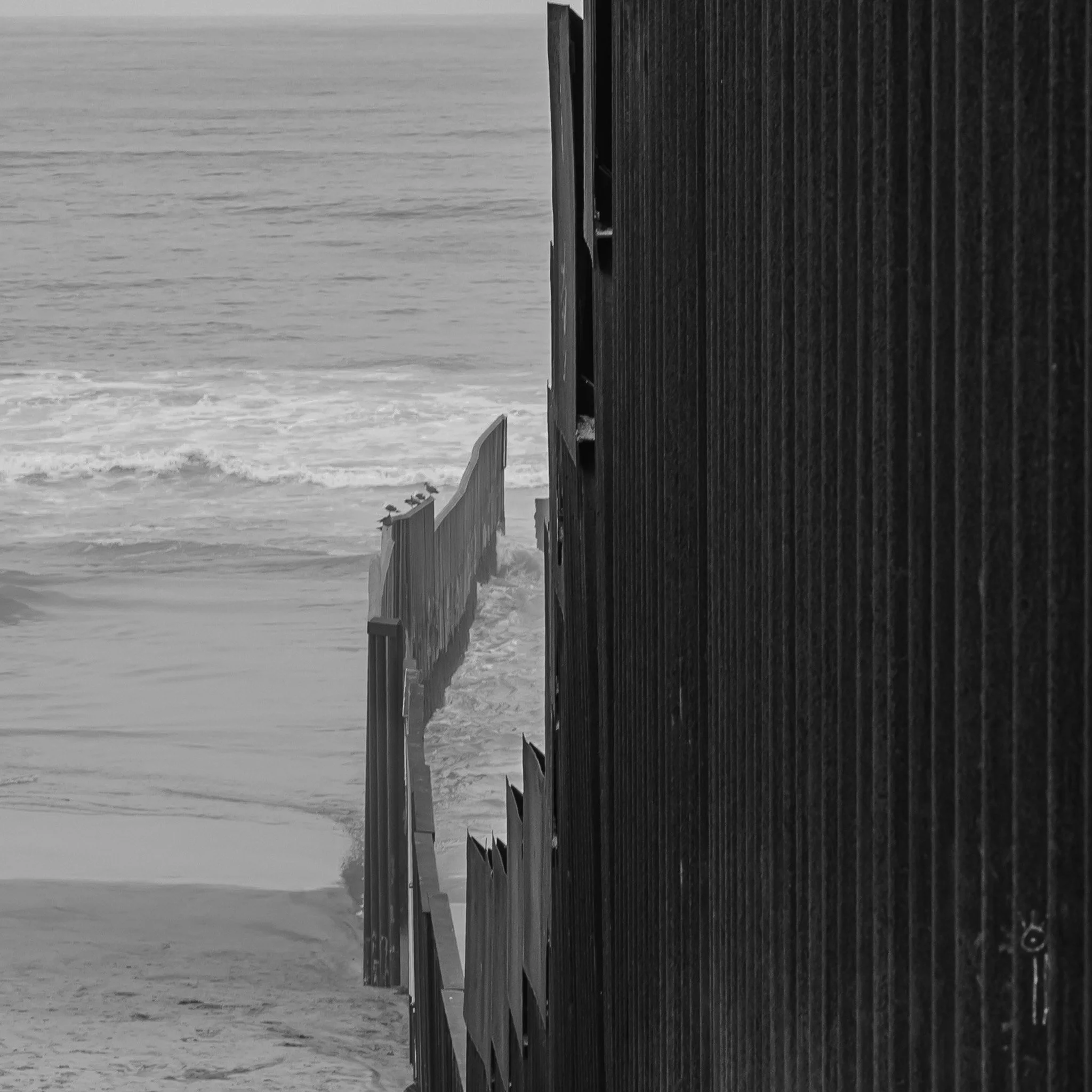 Black and white photo of a metal fence separating the sandy beach from the ocean waves.
