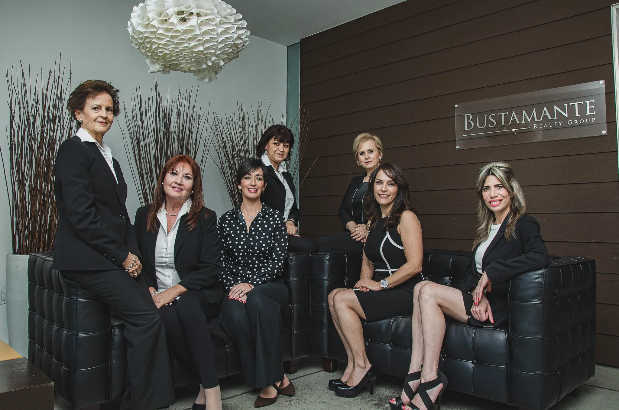 Group of seven women in business attire sitting and standing in office lobby with a sign that reads 'Bustamante Realty Group'.