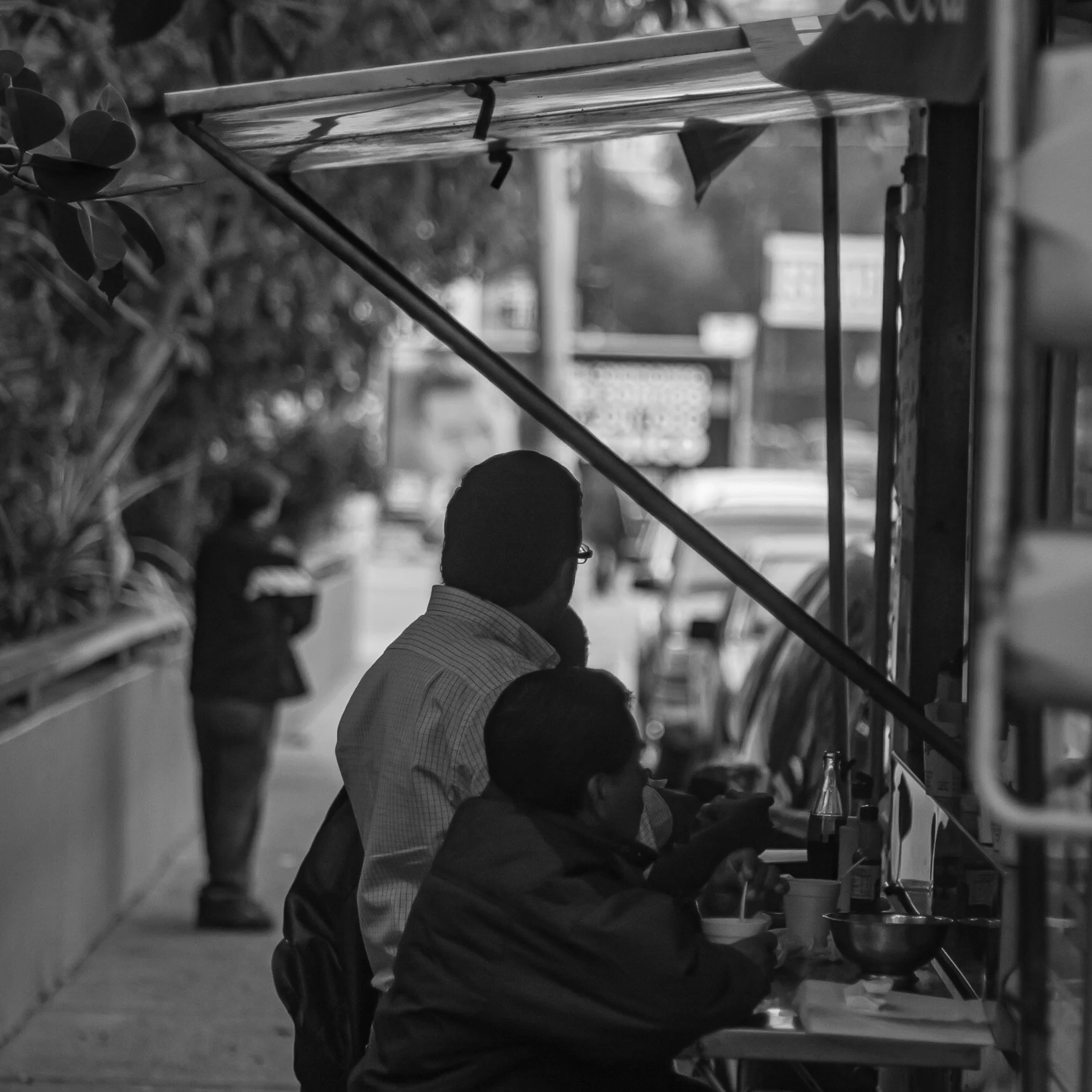Black and white photo of two men sitting at an outdoor counter, eating with chopsticks, with a person in the background walking away.