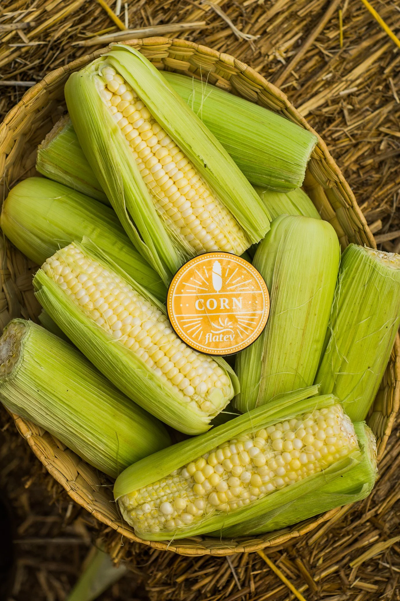 Fresh ears of corn with green husks in a woven basket, some partially husked showing yellow kernels, and a round orange sticker that says 'Corn Flatev'.