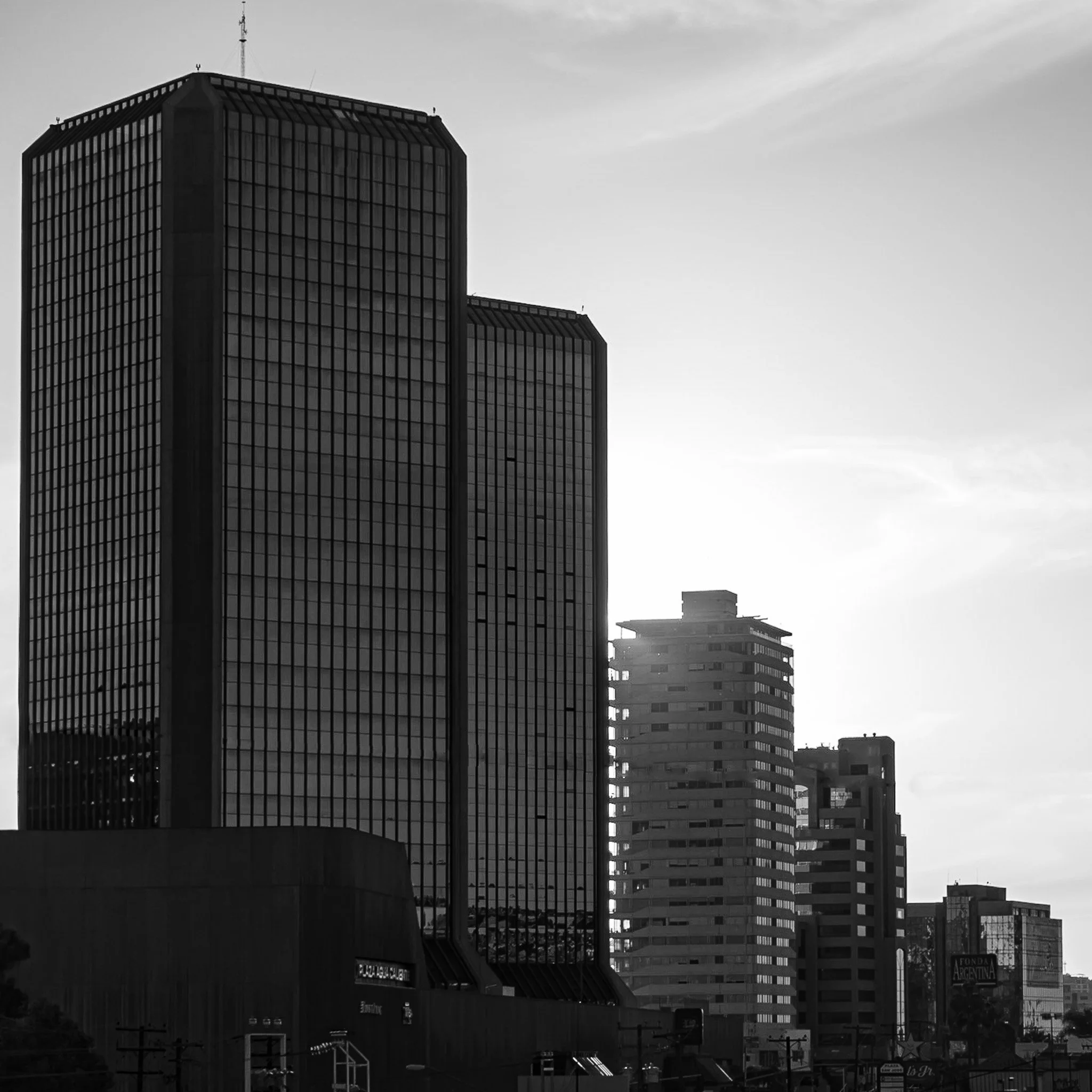 Night cityscape featuring tall modern skyscrapers, some with glass facades reflecting lights, under a cloudy sky in black and white.