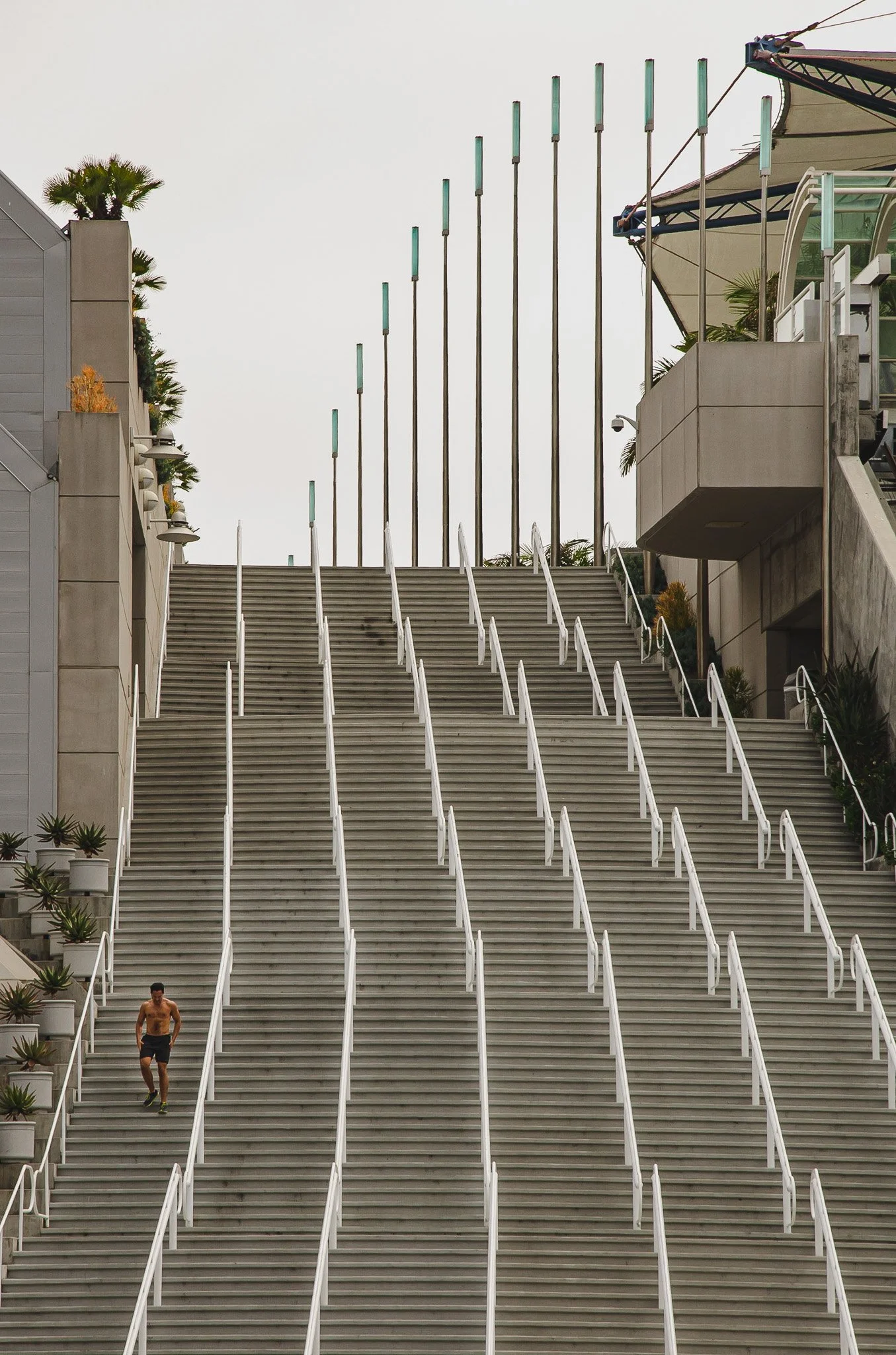 A man running up a long outdoor escalator or staircase with metal handrails on each side, adjacent to modern buildings with potted plants, under a cloudy sky.