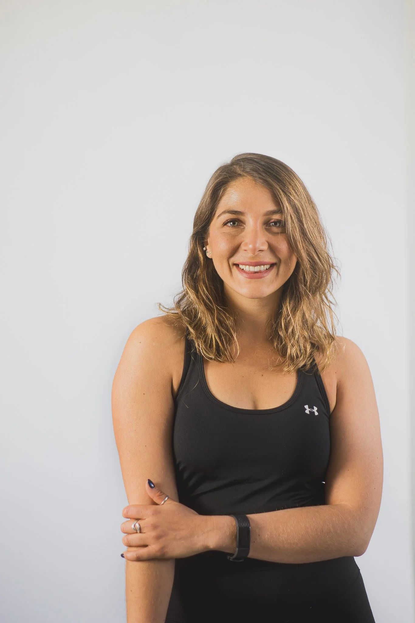A woman with shoulder-length wavy brown hair smiling, wearing a black athletic tank top and a fitness tracker, standing against a plain white background.