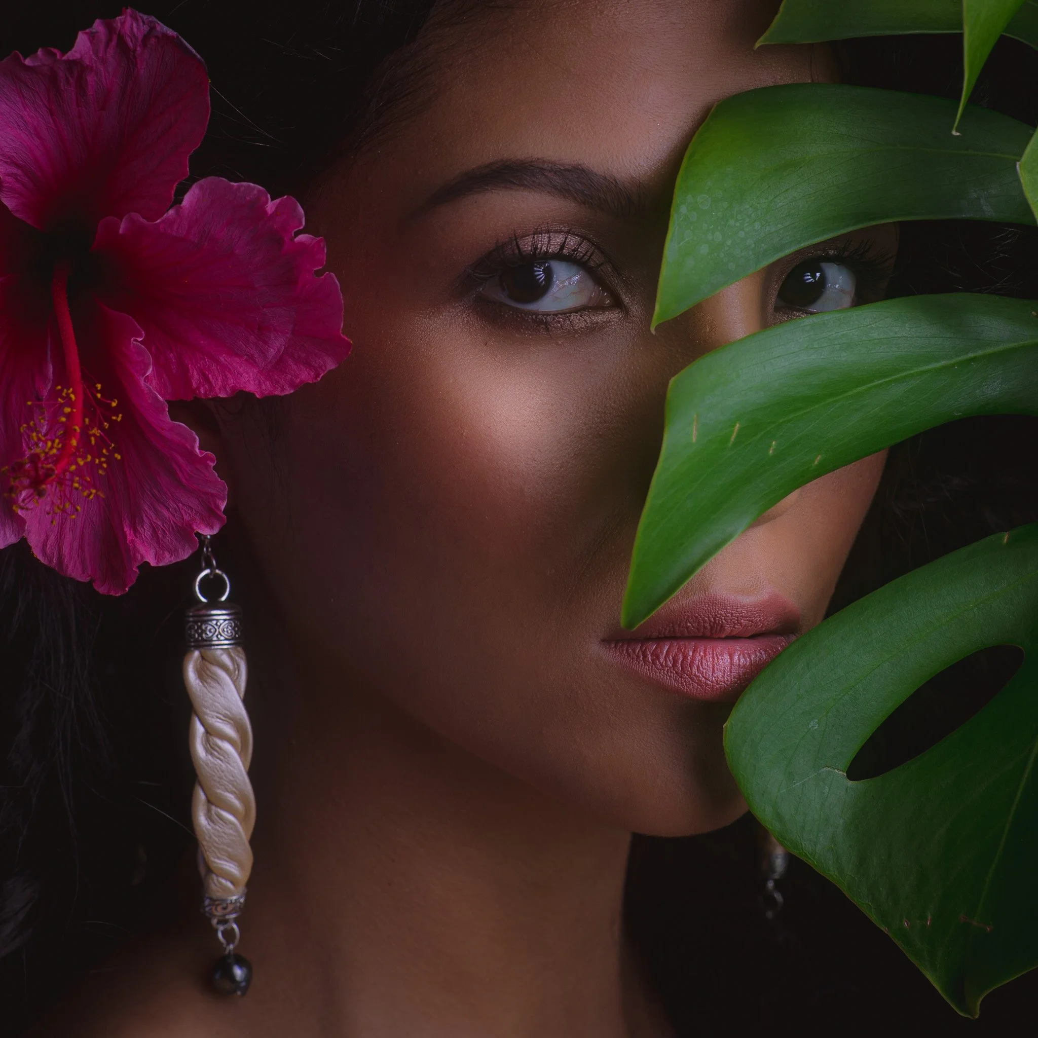 A woman with dark hair and makeup partially obscured by large green leaves and a pink flower, wearing a decorative earring.