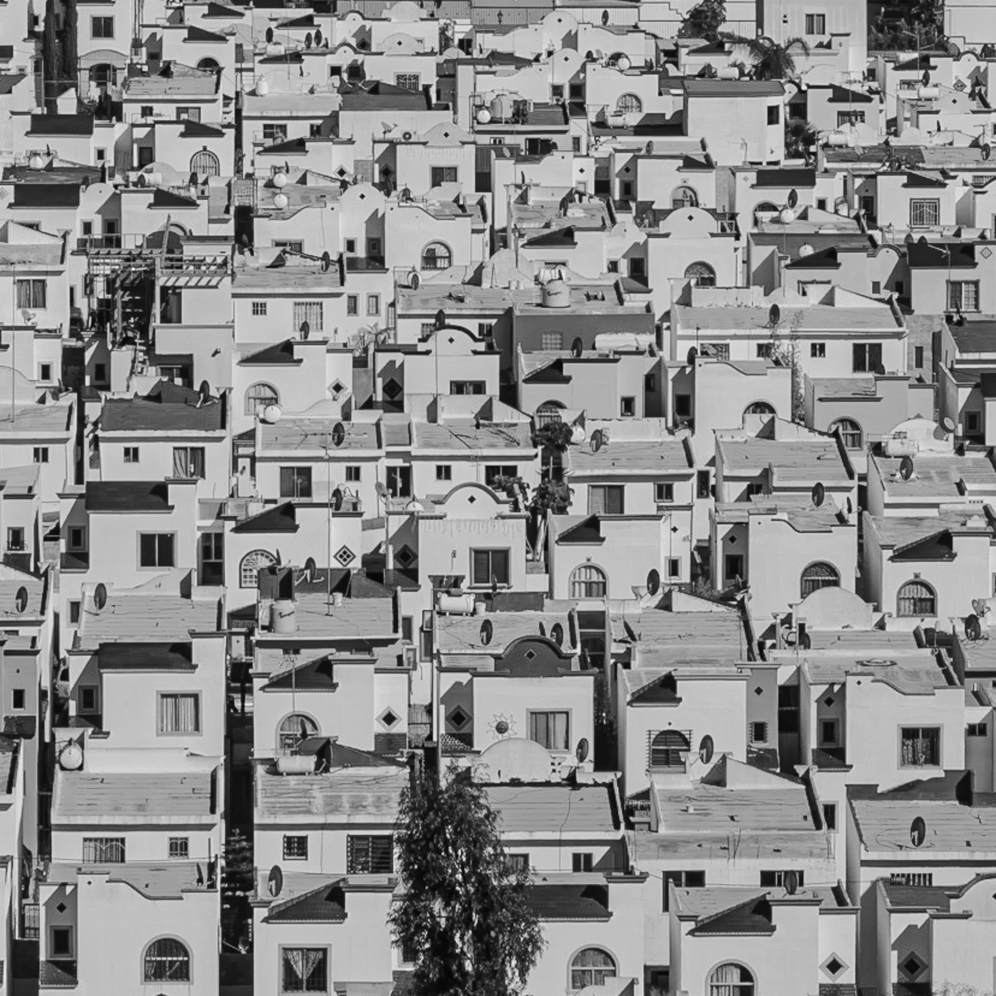 A densely packed neighborhood of closely-built houses with flat roofs, satellite dishes, and small windows, seen from an aerial view in black and white.