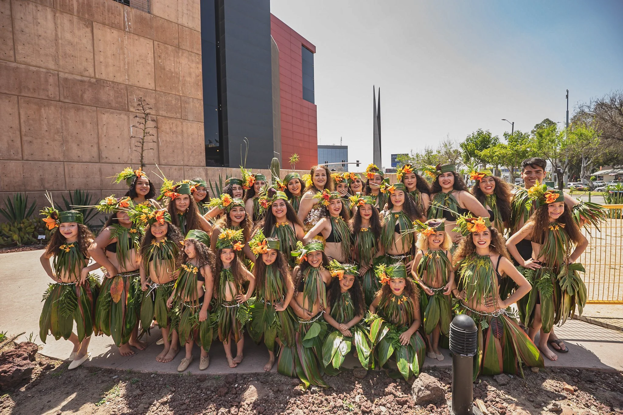 A group of women and young girls dressed in traditional hula costumes made of large green leaves with floral headpieces, gathering outdoors in front of a modern building with a clear blue sky.