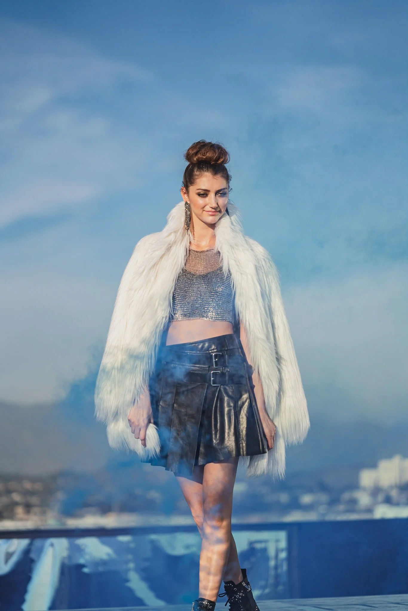 Fashion model walking outdoors on a city rooftop during daytime, wearing a white faux fur coat draped over her shoulders, a metallic crop top, and a black leather skirt with buckle details, with smoke around her legs and a city skyline in the backgro