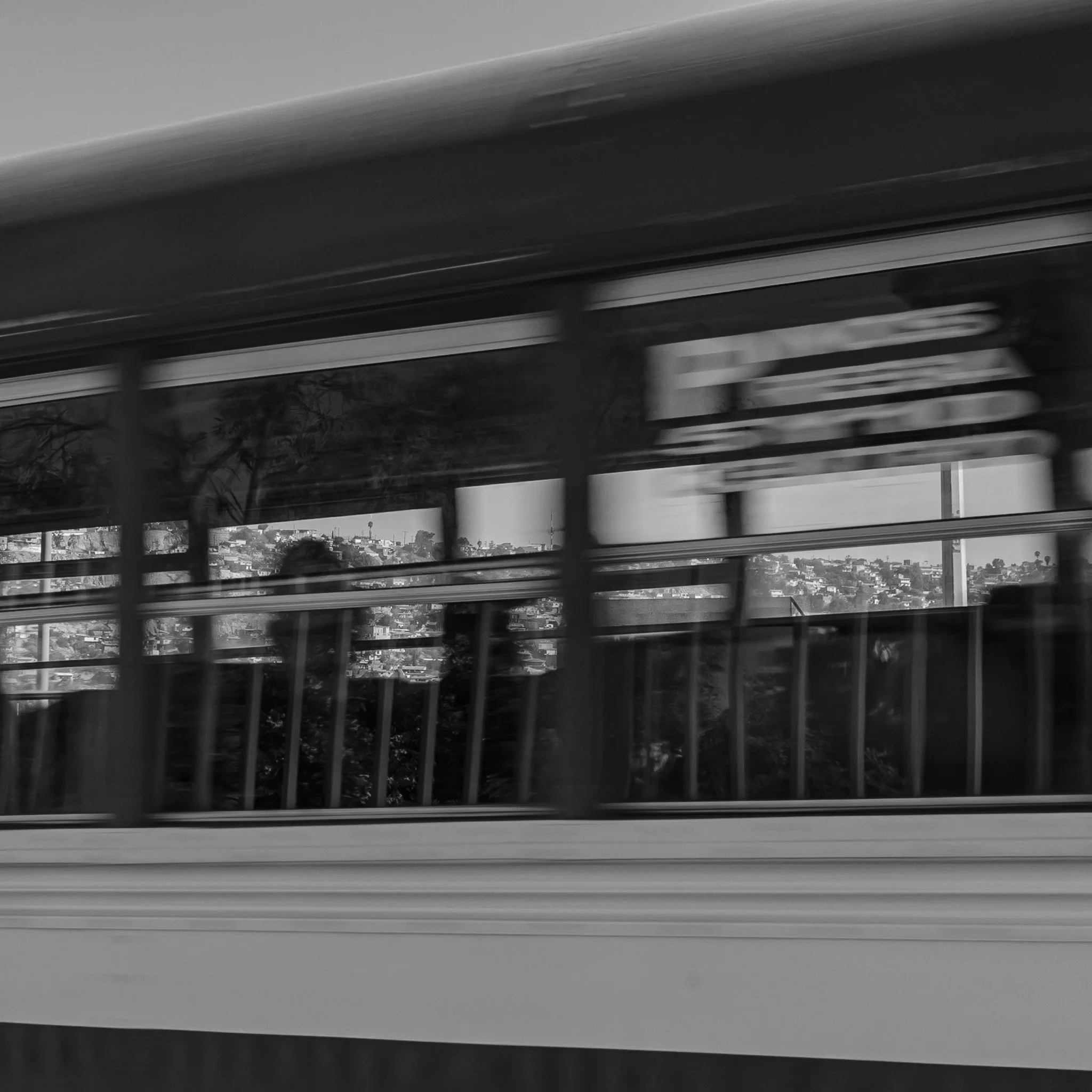 A black and white photo showing a moving train window with a blurred landscape outside, including houses on a hillside.
