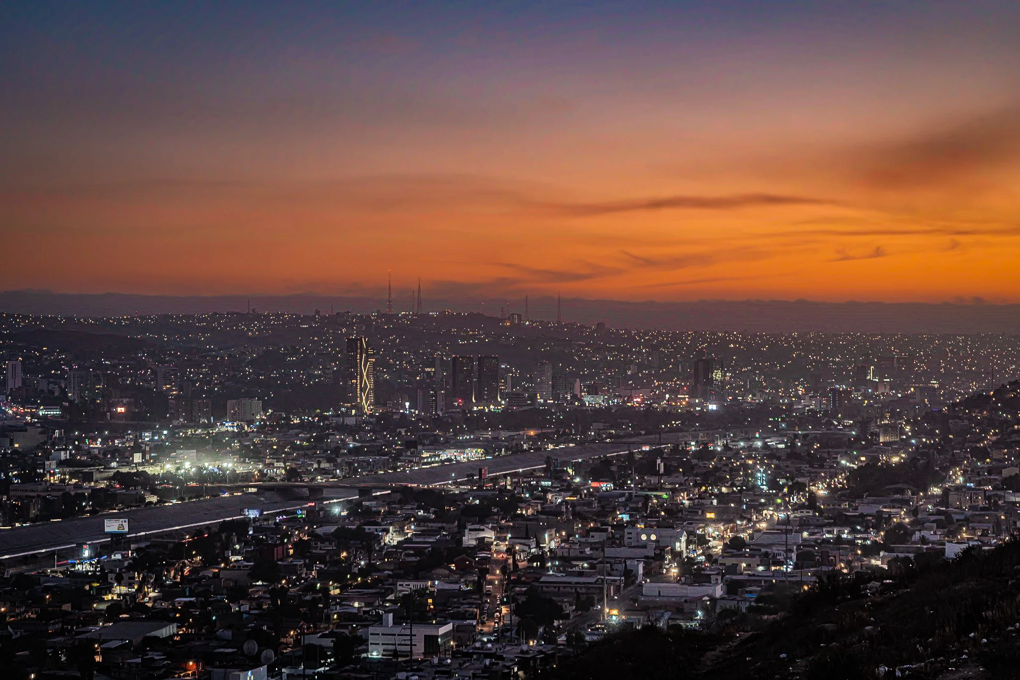 View of a city at sunset with buildings and lights, and a colorful sky with orange, pink, and purple hues.