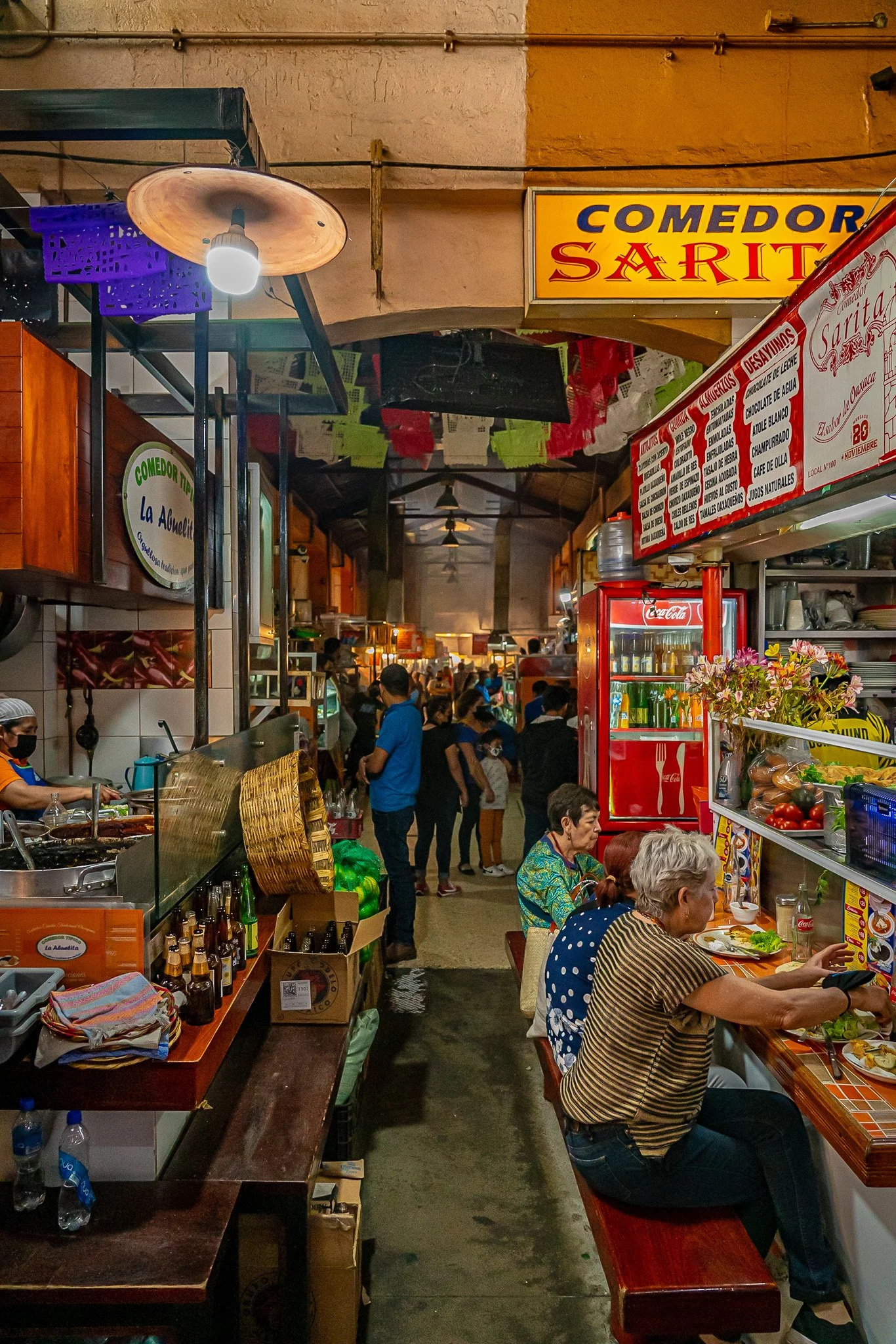 Inside a busy Mexican food market with food stalls, colorful signs, and customers dining and shopping.