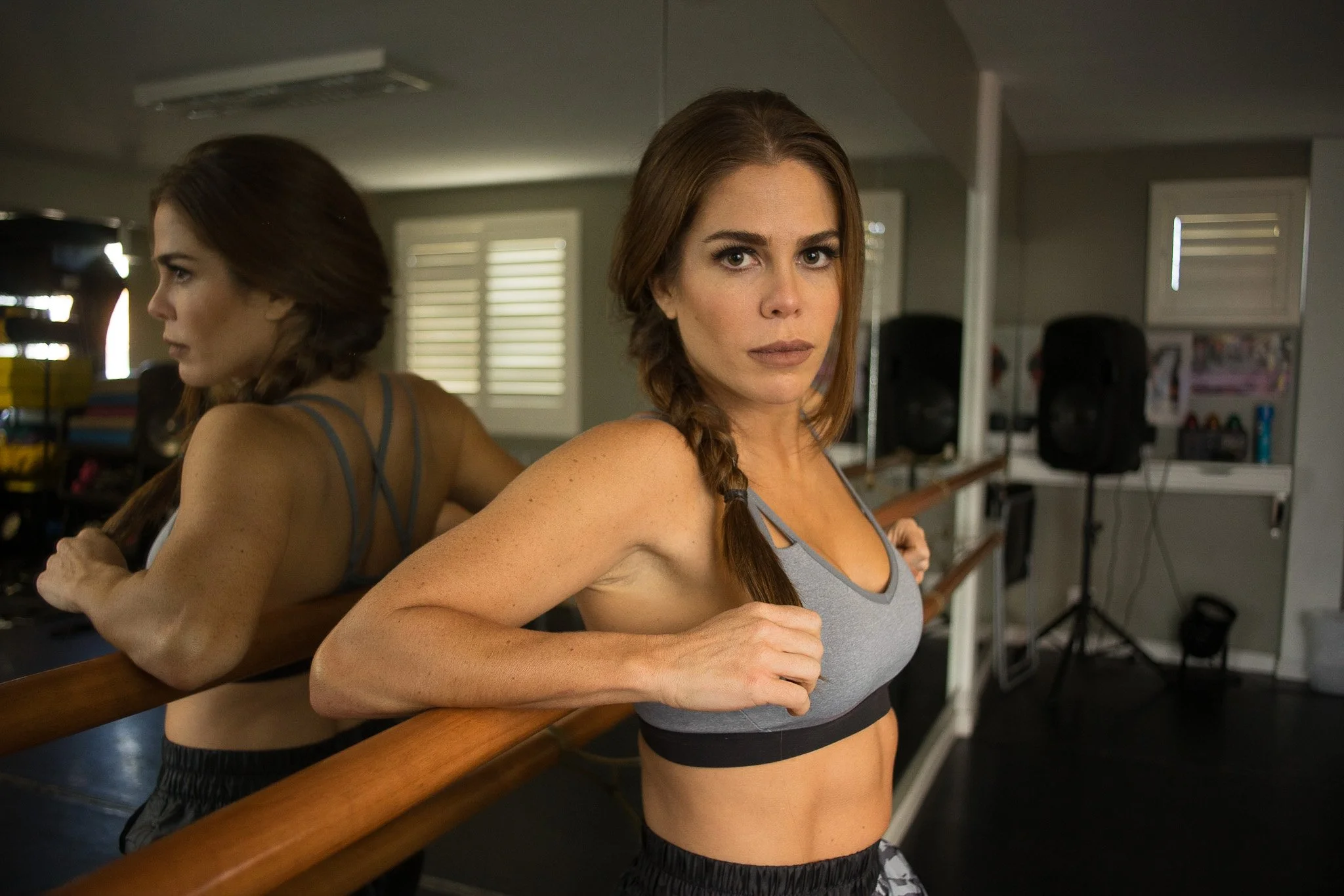 A woman with braided hair and fitness sportswear leaning on a ballet barre in a gym studio with mirrors.