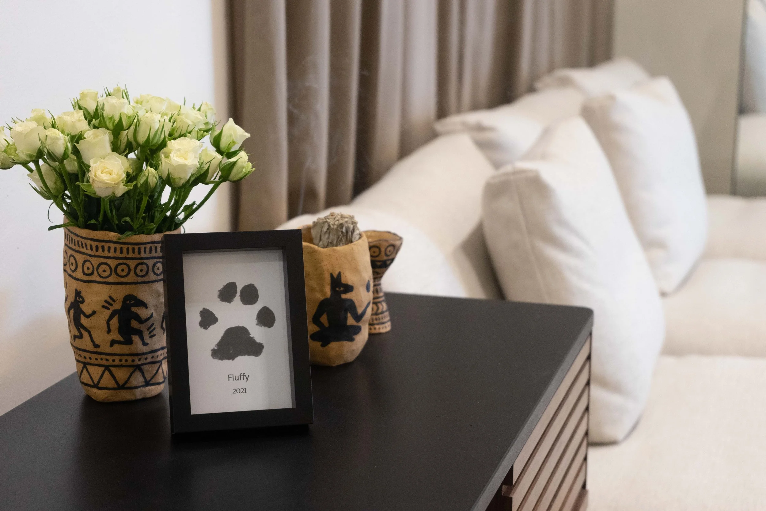 A black side table with a framed paper showing a black paw print, a vase of white roses, and two small decorated ceramic pots, all in a room with white sofa cushions and beige curtains in the background.