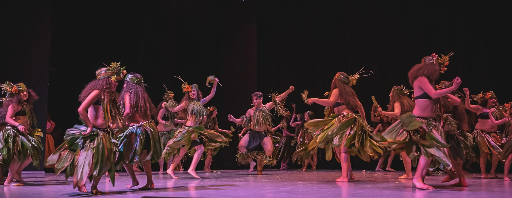 Group of dancers performing on stage, dressed in traditional Polynesian costumes made of leaves, some wearing headdresses, dancing in a circle.