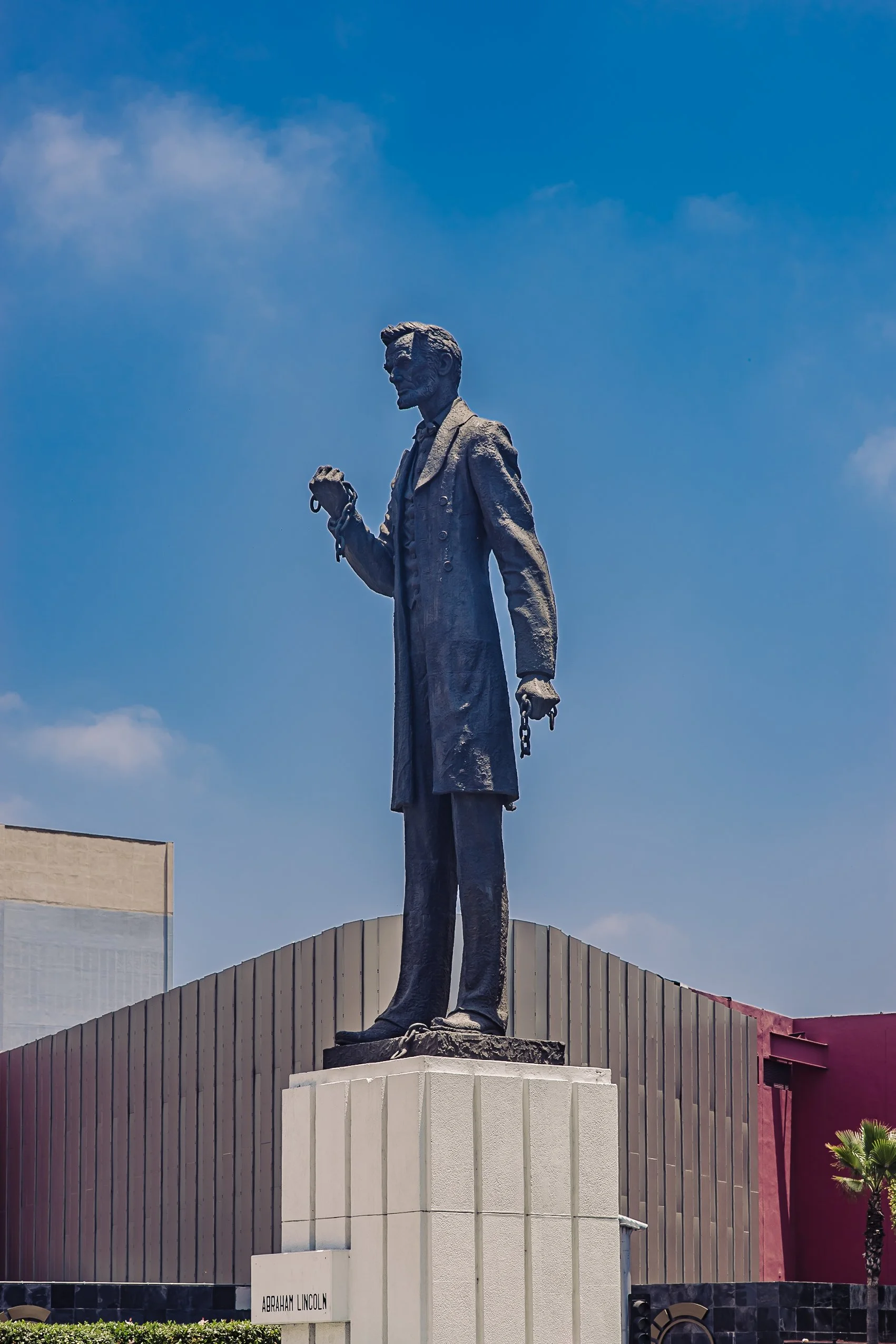 Statue of Abraham Lincoln holding a pair of glasses in one hand, with the other hand chained, on a white pedestal in front of a modern building against a blue sky.