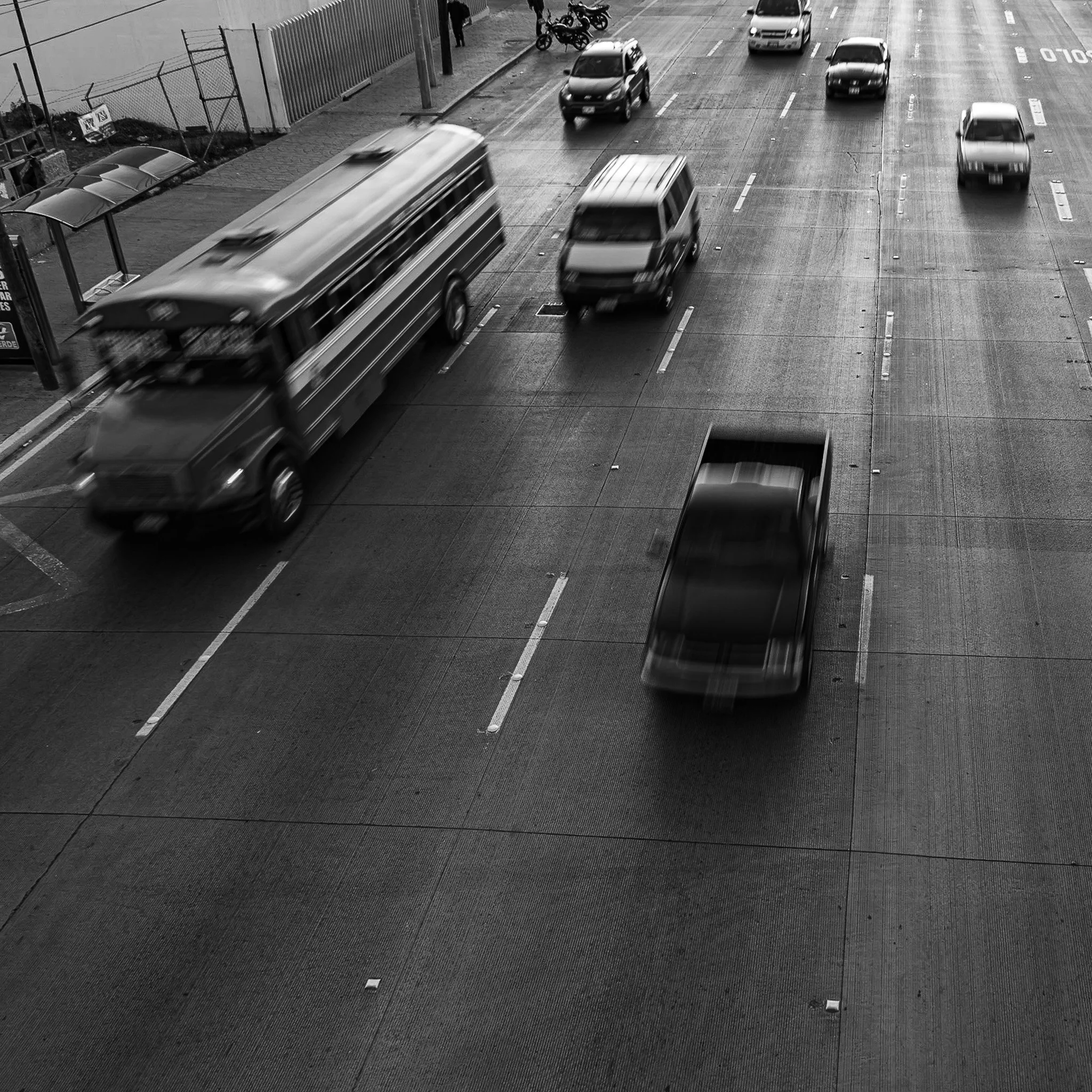 Black and white photo of a busy city street with several moving cars, some parked bikes, and a bus. The cars appear blurred due to motion. There is a fence and a small bus stop on the left side, and the street has visible Lane markings.