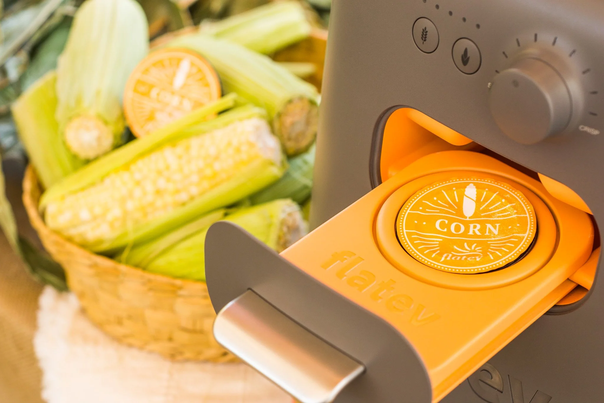 Food dehydrator with a label reading 'CORN' from a jar of dried corn inside, next to a basket of fresh corn on the cob and a sliced orange.