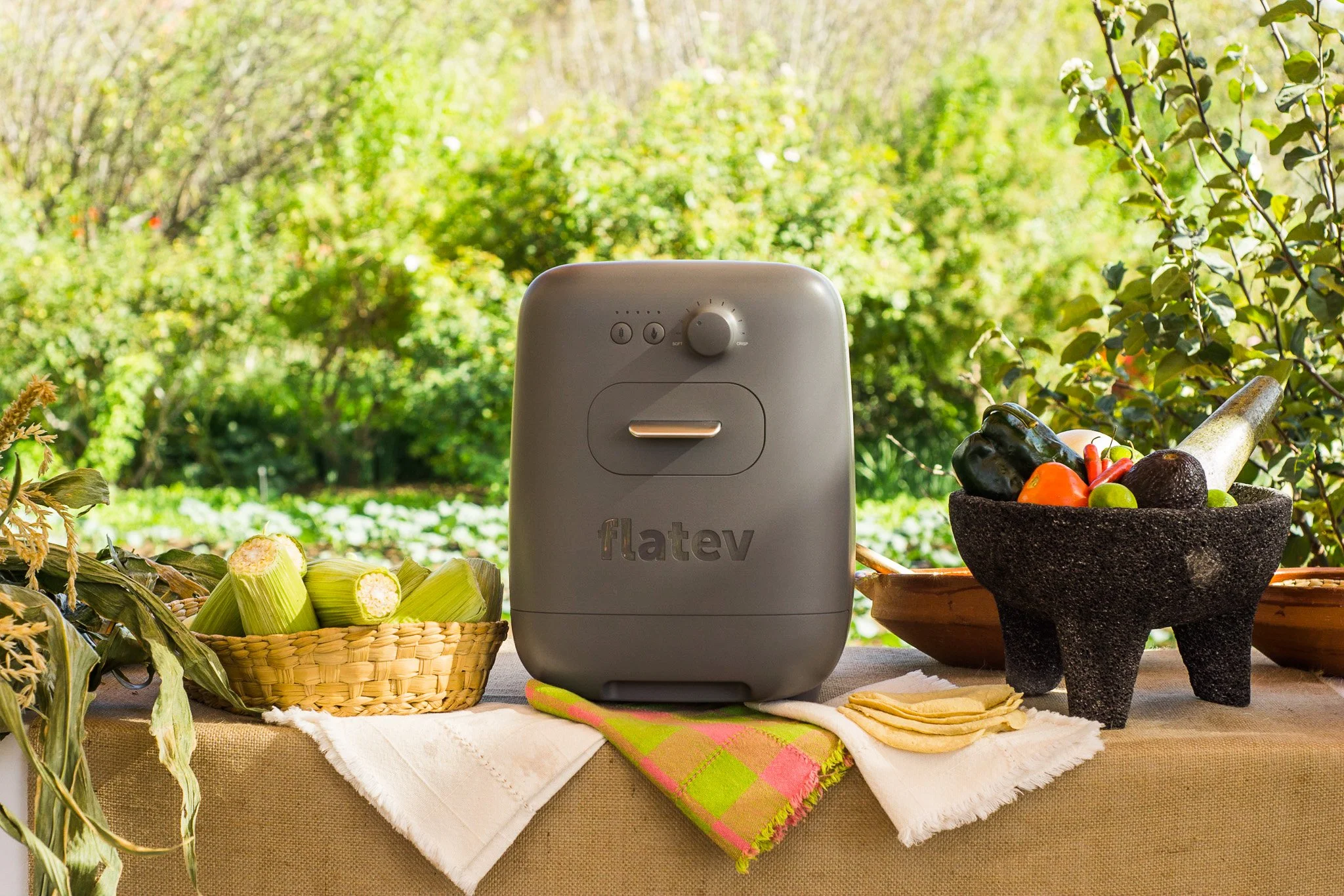 A countertop with fresh vegetables, a charcoal-colored molcajete filled with vegetables, and a grey Flatev automatic tortilla maker, set outdoors with greenery in the background.
