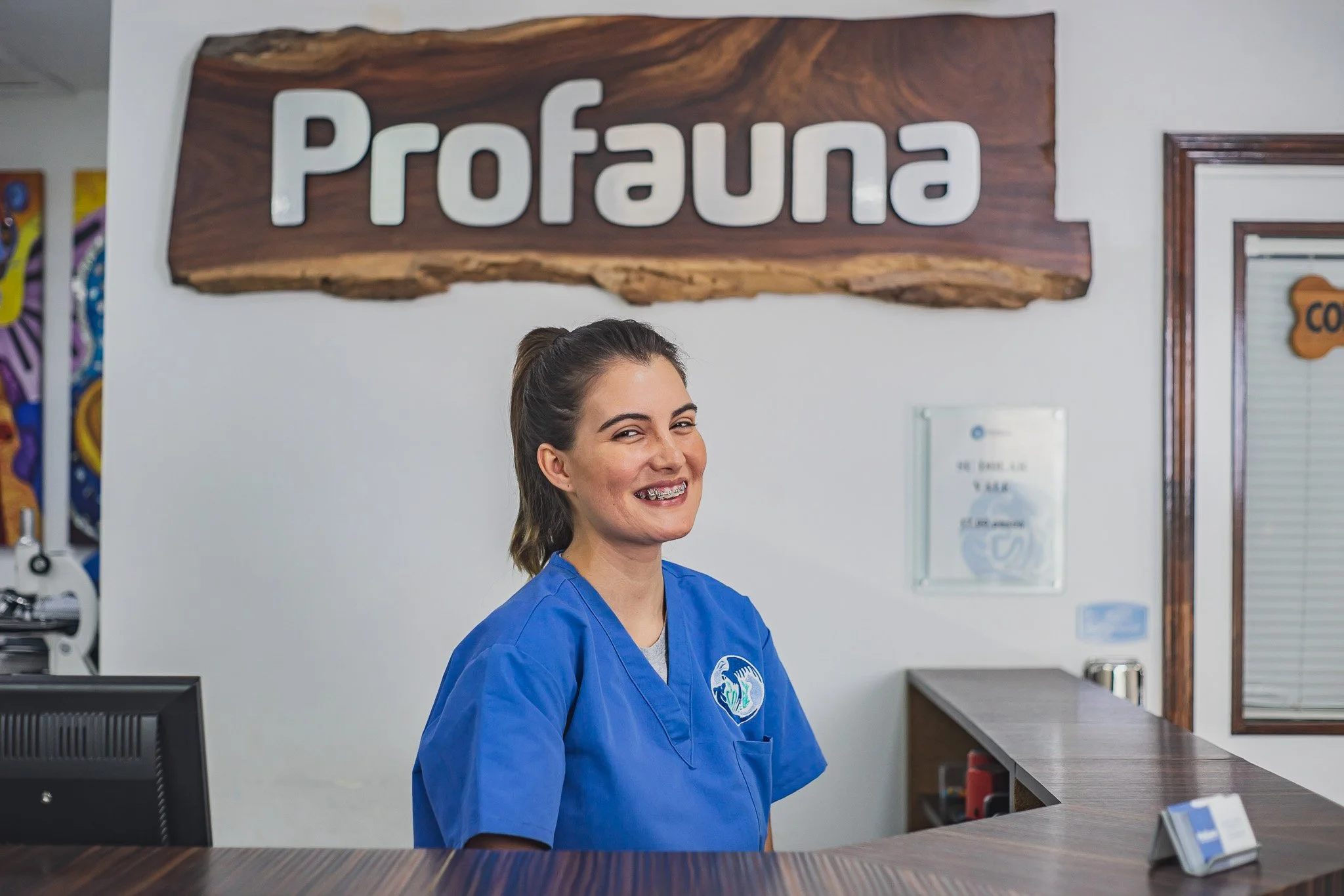 A smiling woman with braces wearing a blue uniform behind a reception desk at a place called Profuna.