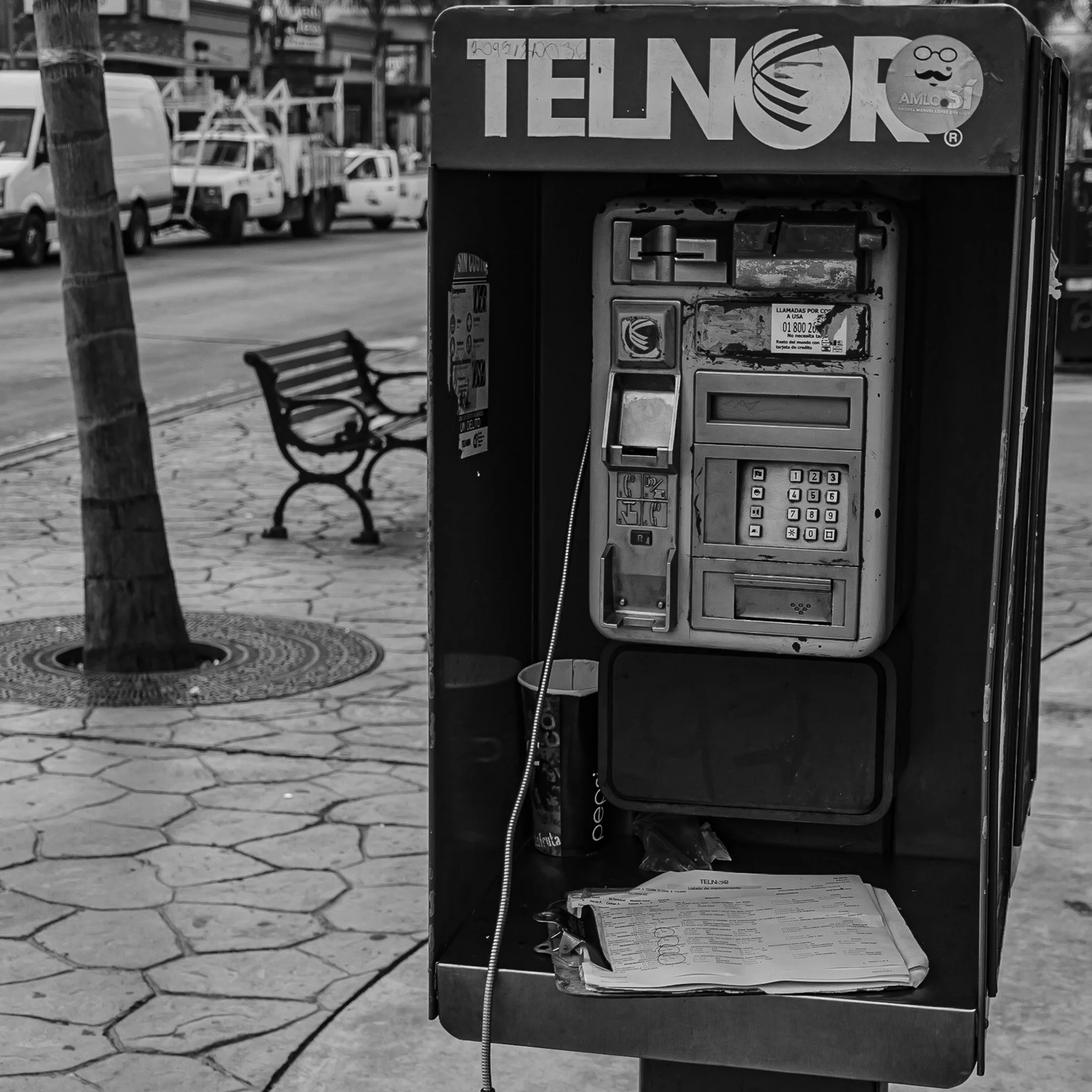 An old payphone booth on a sidewalk with a phonebook resting at the bottom and a trash can beside it. The booth has the word TELNOR at the top, with some stickers and writing, and a bench with benches and parked cars in the background.