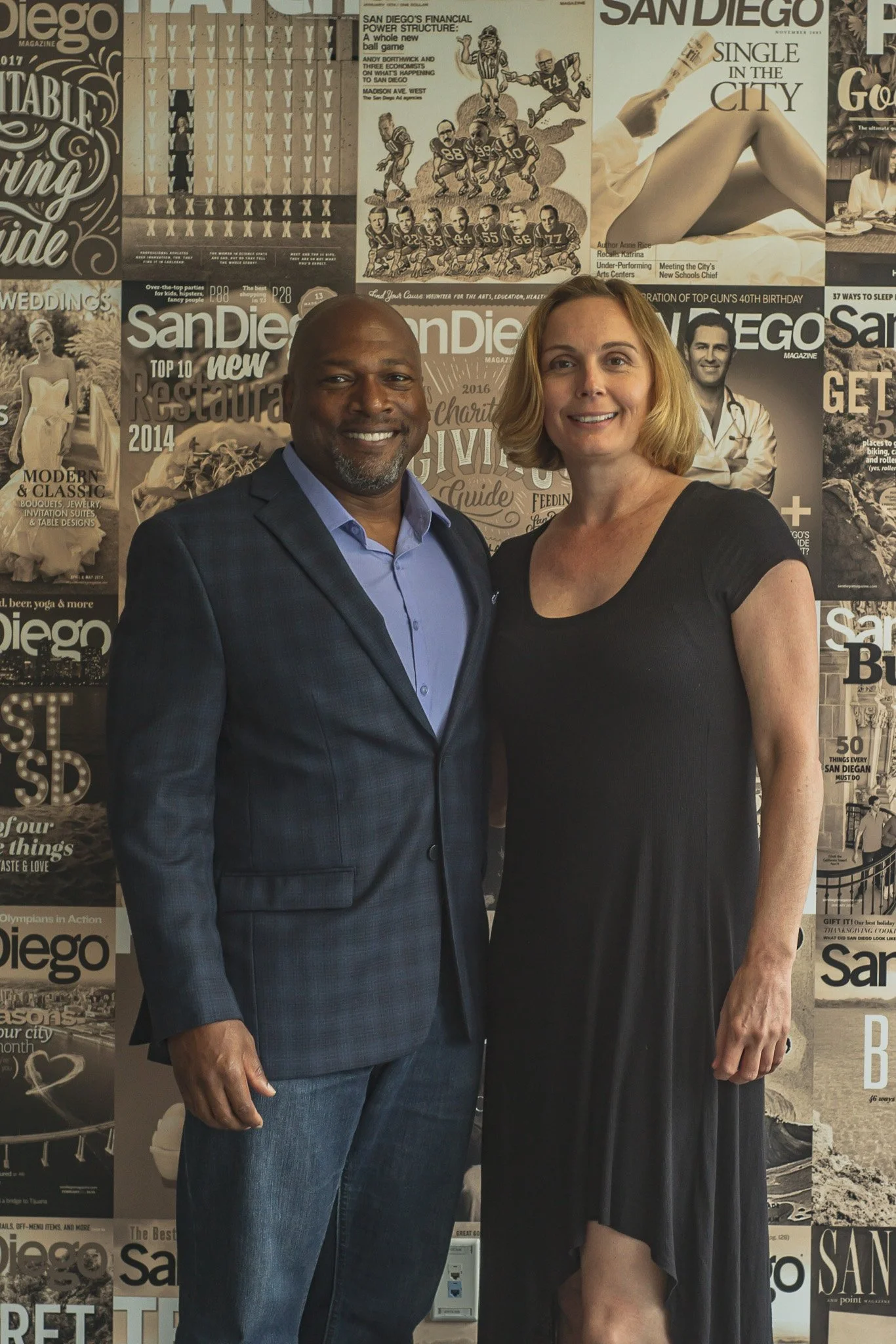 A man and woman standing side by side in front of a collage of magazine covers, smiling at the camera. The man is dressed in a navy blue checked blazer and blue shirt, and the woman is wearing a black dress with short sleeves.