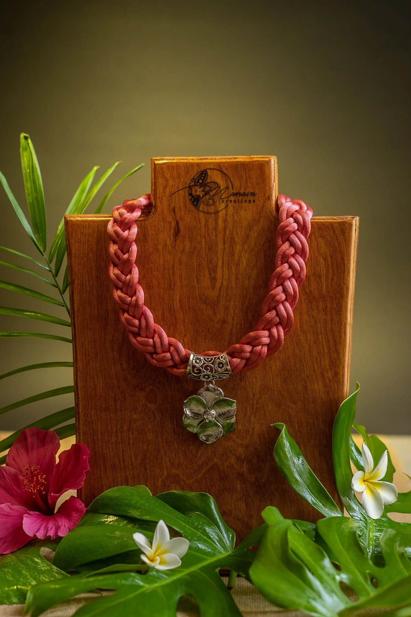 A pink braided necklace with a flower-shaped pendant on a wooden display stand surrounded by tropical flowers and green leaves.