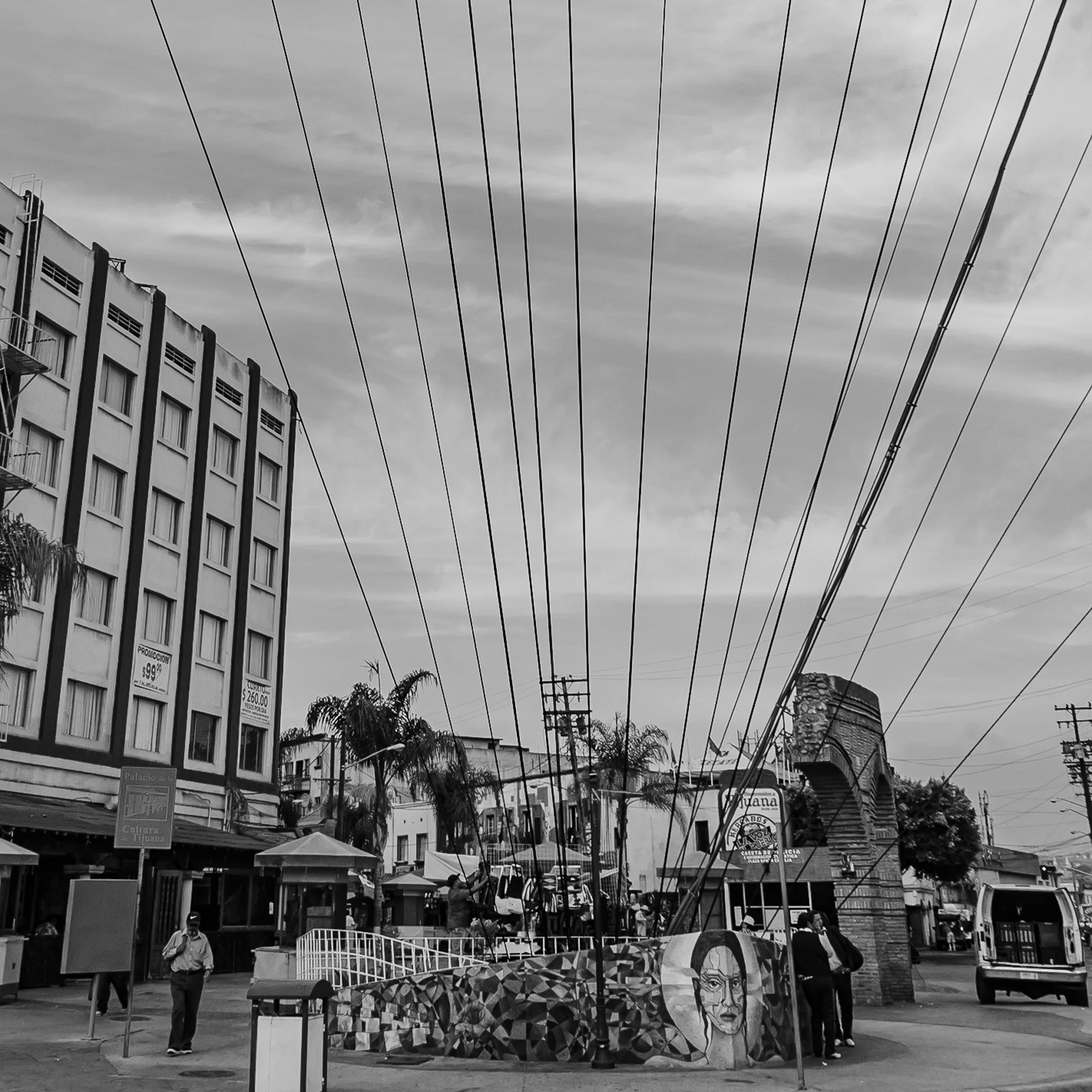 A black and white photo of an urban street scene with a small roundabout featuring a mural of a woman's face, surrounded by several people, palm trees, a bus, and buildings with utility poles and power lines overhead.