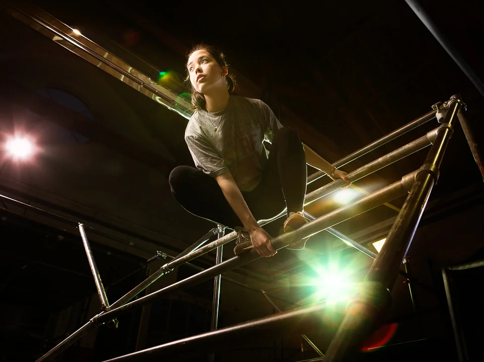 I didn't have much of a plan going into this parkour shot, other than I wanted to heavily light it, and darken the (busy) backgrounds both out of necessity and aiming for drama. But to get an action shot (at least, the ones I'd have liked to capture) would require more precise lighting than there was time for. So it became something very static, rather defeating what the sport is about! When she posed, looking at camera, it just didn't work at all. We came up with the idea that she's readying herself - well, planning, at least, by her expression - to jump, and it turns out that potential energy isn't too bad a tradeoff for kinetic.