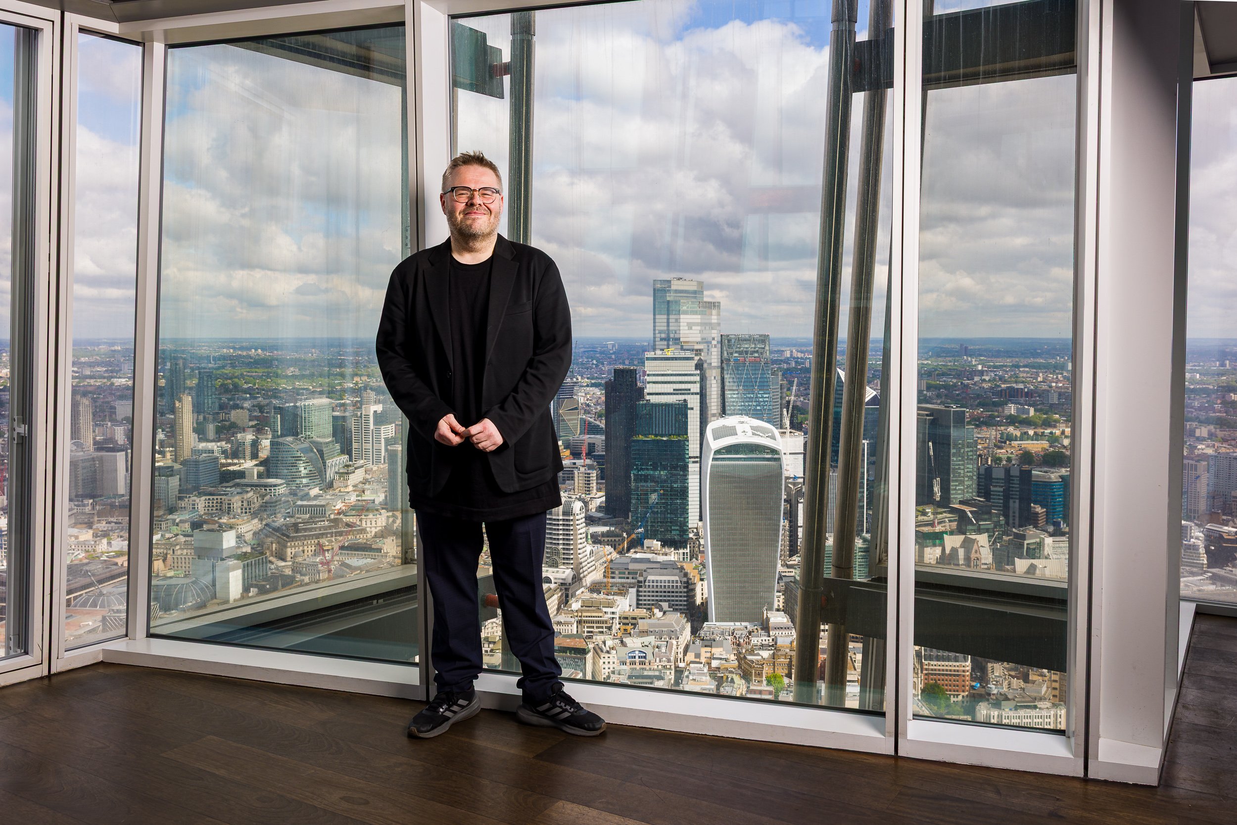 Man in a dark suit stands in the Shard, with the City of London in the background