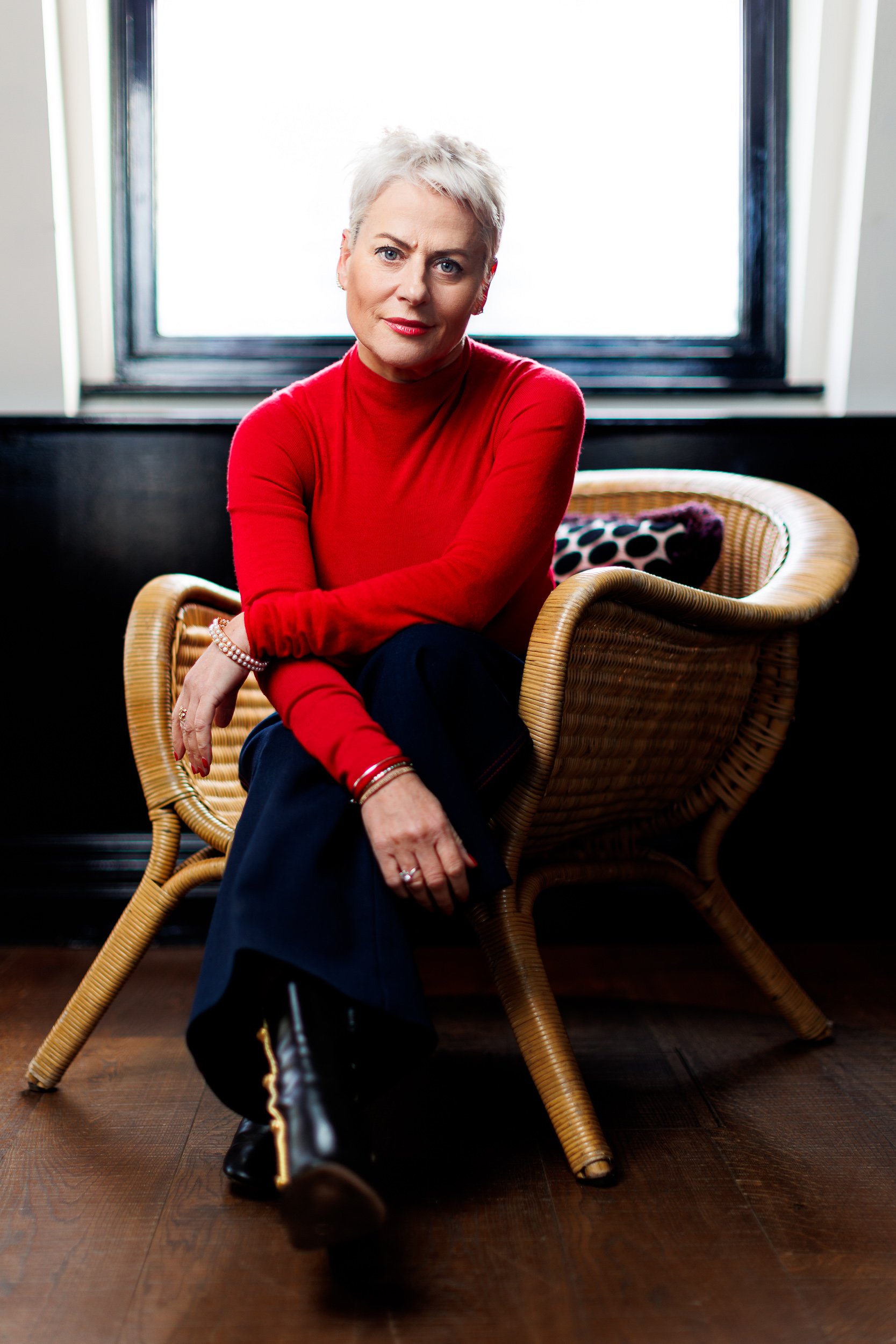 A woman in a red jumper sits in a chair and looks at camera