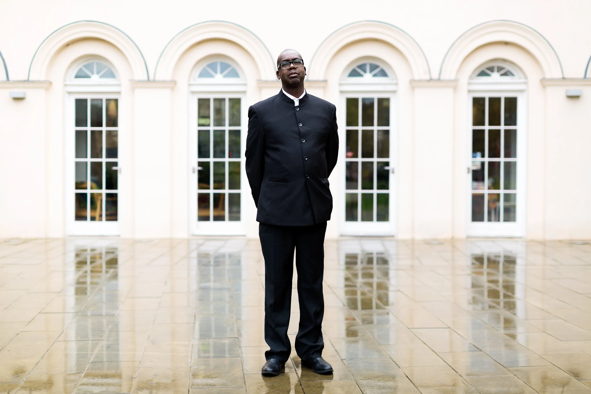 A man in a dark suit stands in front of an old building in the rain