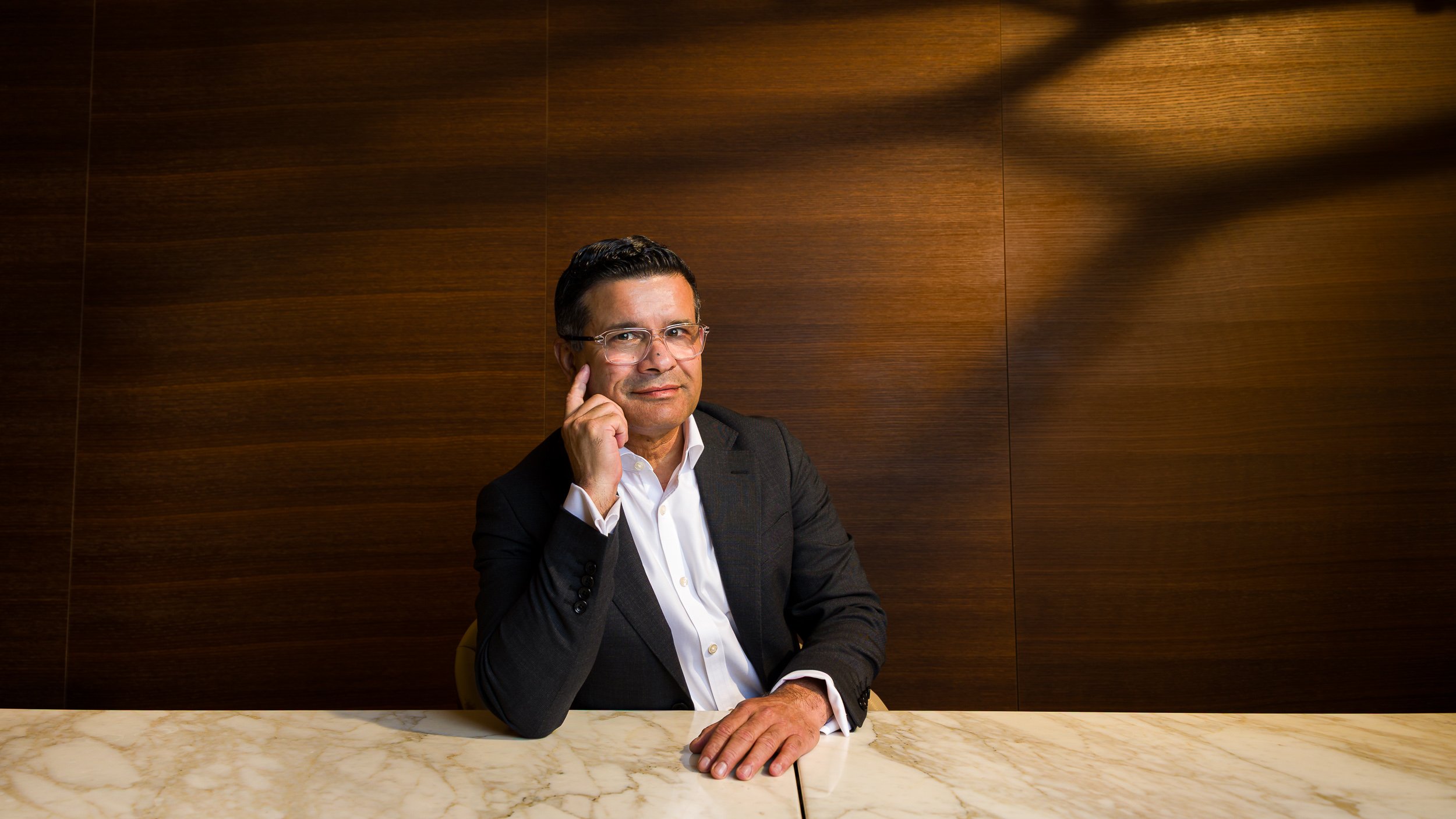 A man in a suit sits at a table and looks at camera