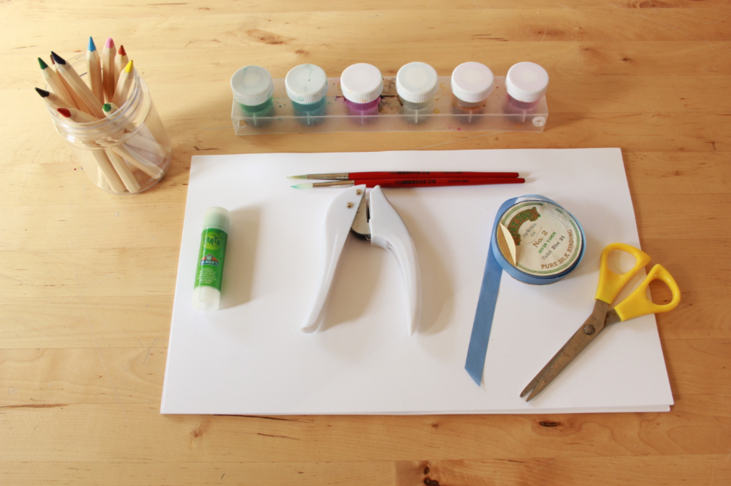 Gather supplies, paper, glue stick or stapler, hole punch, string, scissors, pencils, watercolor.