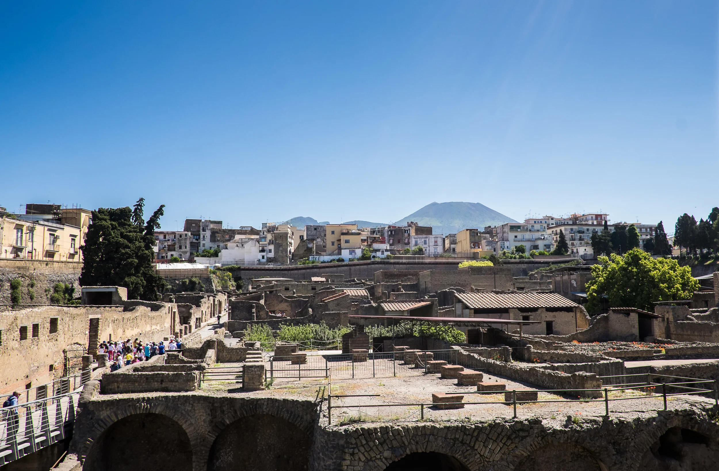 In 79 AD, this city was buried with volcanic mud from Mount Vesuvius -- seen in the background -- similarly to how Pompeii was buried in volcanic ash. The mud did a superior job of preserving this site; roughly 20% of the city has been excavated. The rest cannot, due to the modern-day city of Ecolani which sits on top of it, seen in the middle-ground. 