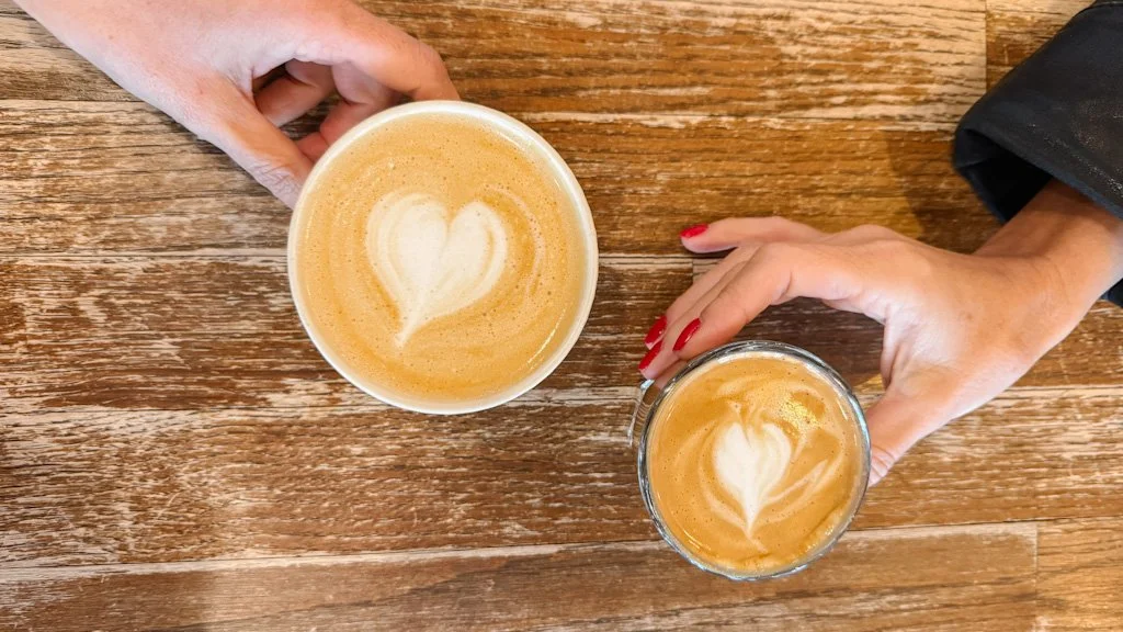 Deux mains tiennent chacune une tasse de café présentant une mousse en forme de coeur sur un table en bois