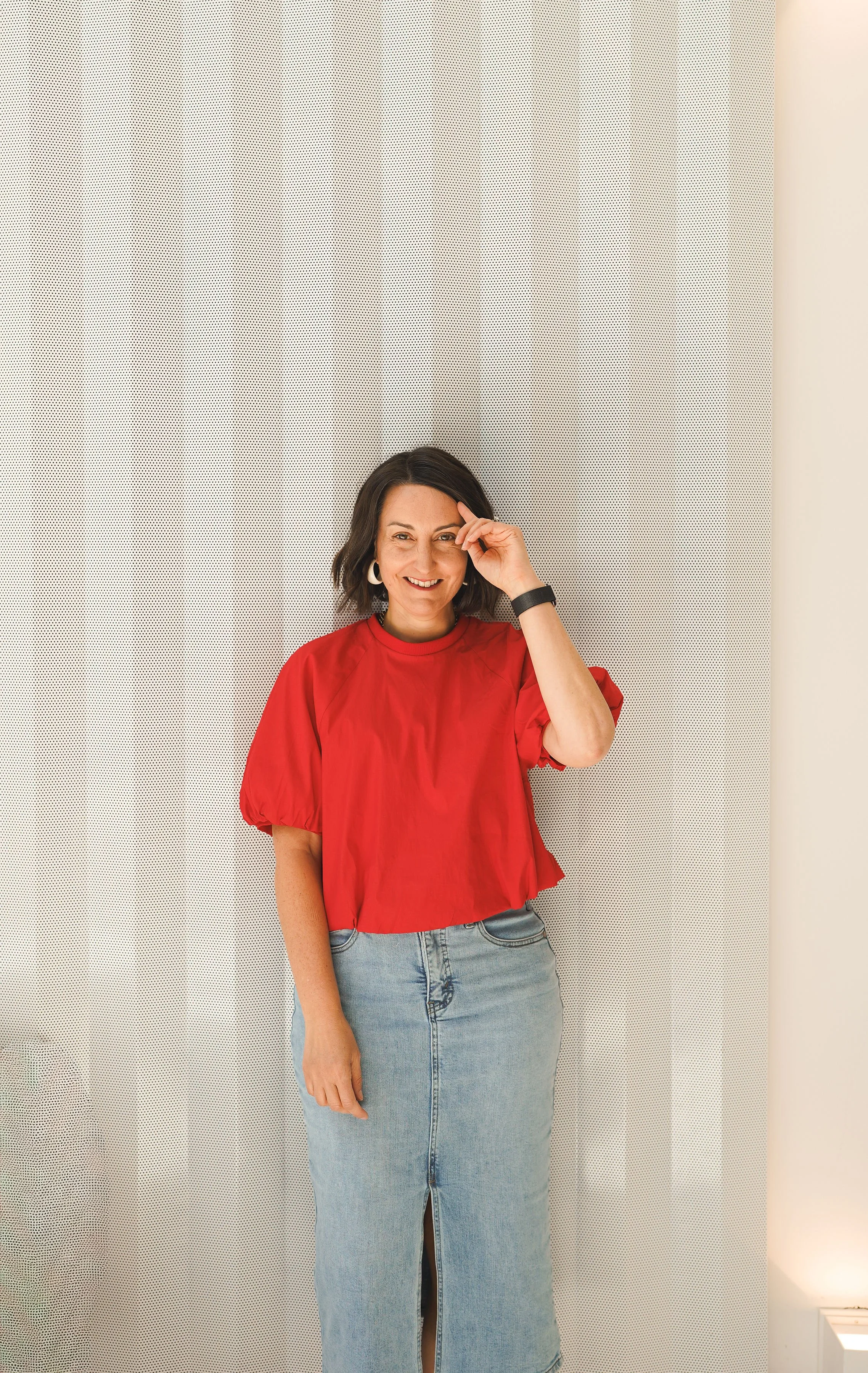Portrait de Jaime en haut rouge et jupe en denim, souriante devant un rideau