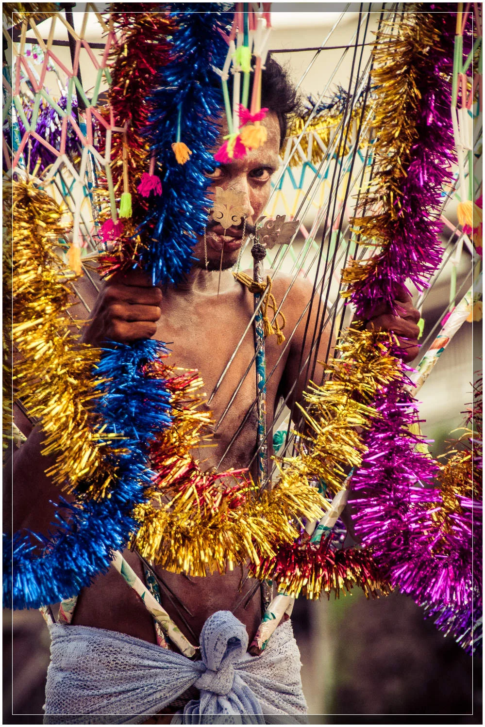 A devotee carrying a vel kavadi (burden) made of metal spears piercing his torso, with a decorated canopy overtop. The tongue is pierced to be a constant reminder of god. © Dustin Main 2013