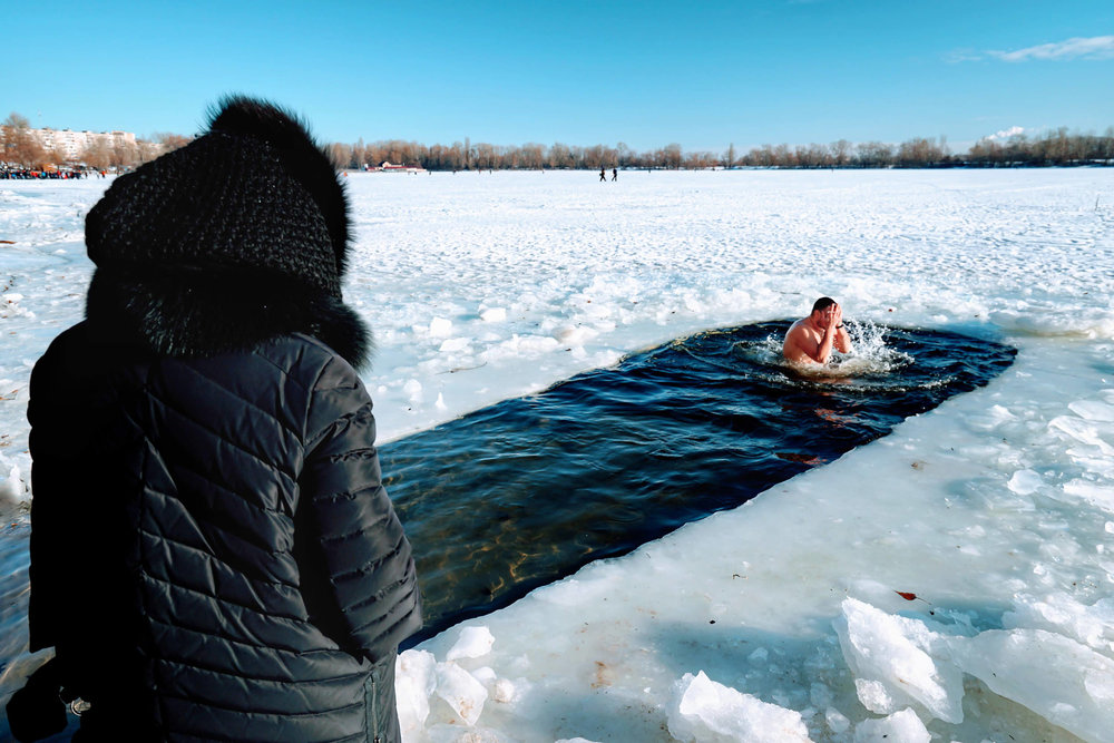  Warmly dressed woman watches a man swim in the frigid waters of the Dnieper river. © Dustin Main 2019 