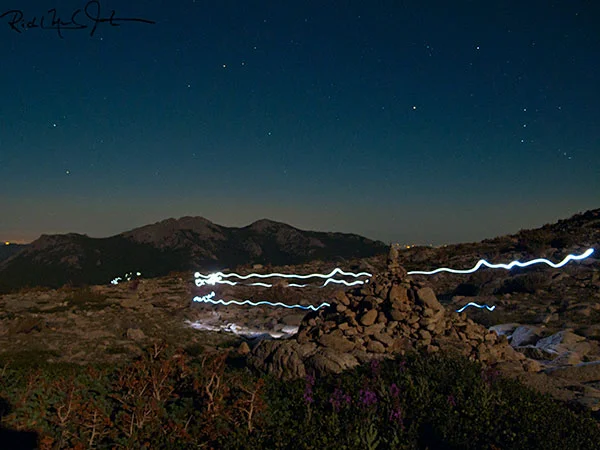 3:04 AM: Headlamps of hikers appear like giant fireflies as they pass a stela marking the route to the summit of Long's Peak. Orion's Belt (three equally-spaced stars in a vertical line) floats above the horizon at right.