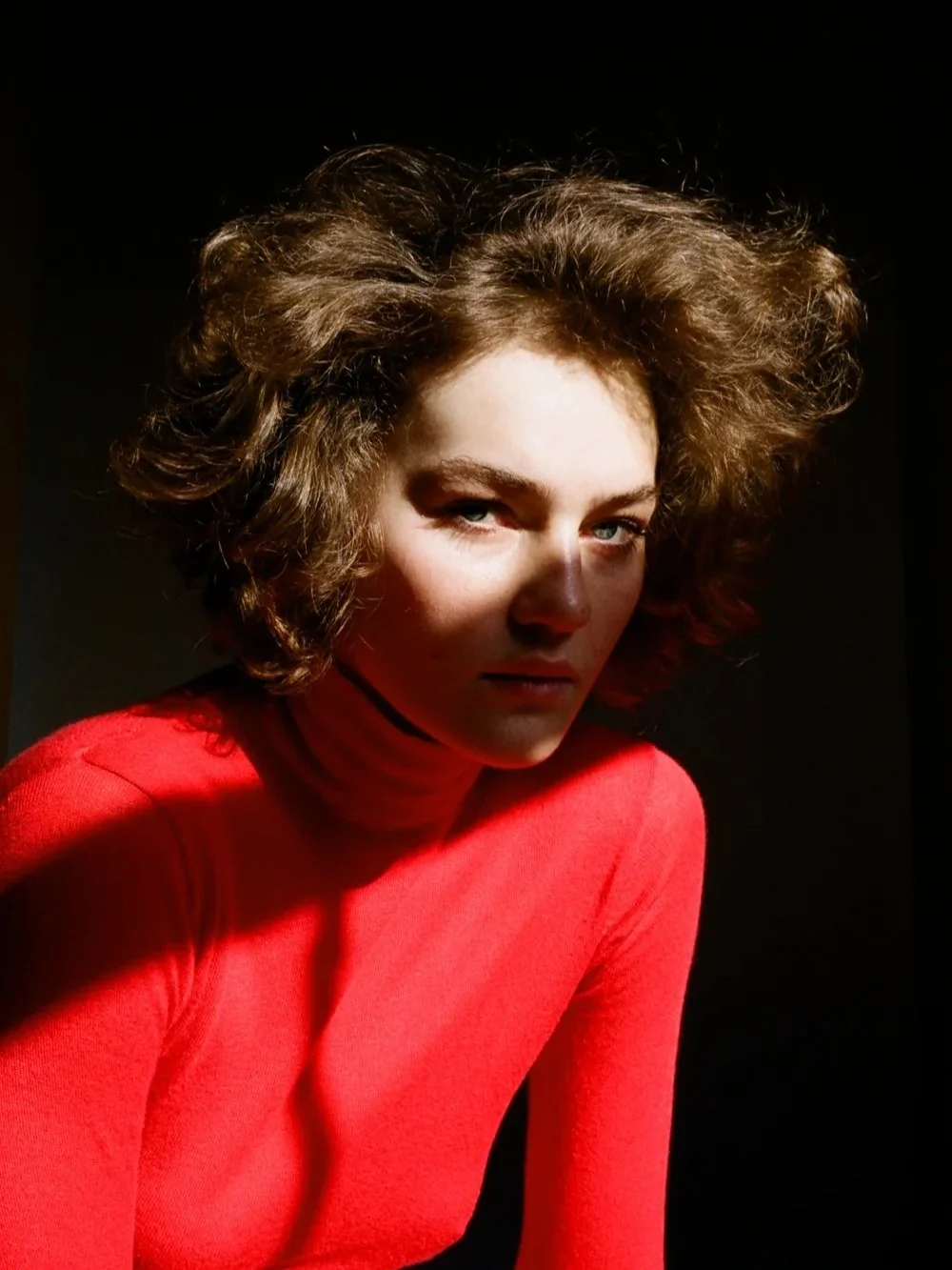 A woman with curly brown hair and blue eyes wearing a bright red turtleneck, looking into the camera with low light and a shadowed background.