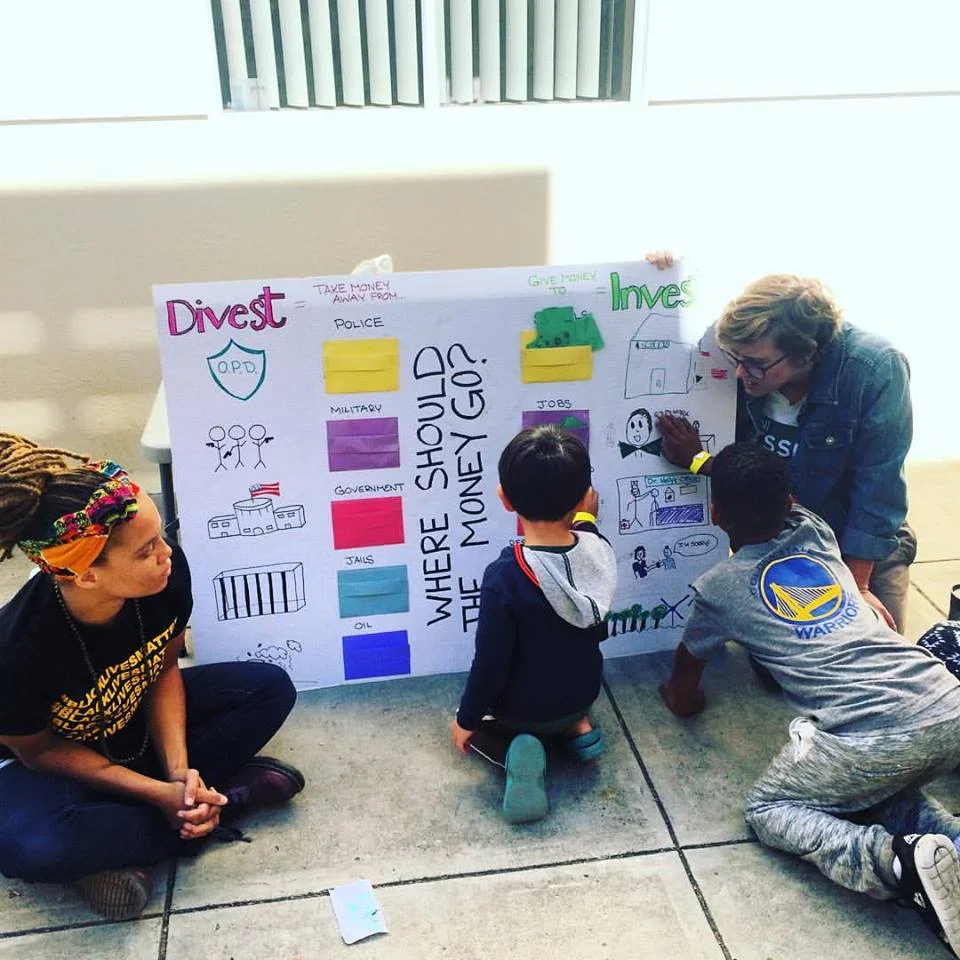 A moment of learning at the Children's Town Hall, as part of the BLM Election Town Hall. At this interactive learning station, children explored the meaning of Divest-Invest, from the Movement for Black Lives Platform. They decided where our money should go by placing dollars into different pockets on the board. The children demand that we invest in schools!